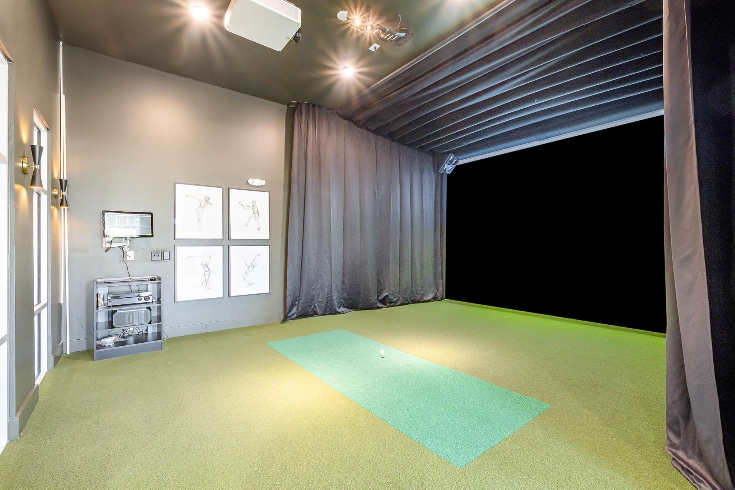 Indoor golf simulator room with green turf flooring, a black curtain for privacy, and framed artwork on the wall. The space is well-lit, with a projector or screen mounted above, and a practice area ready for use.