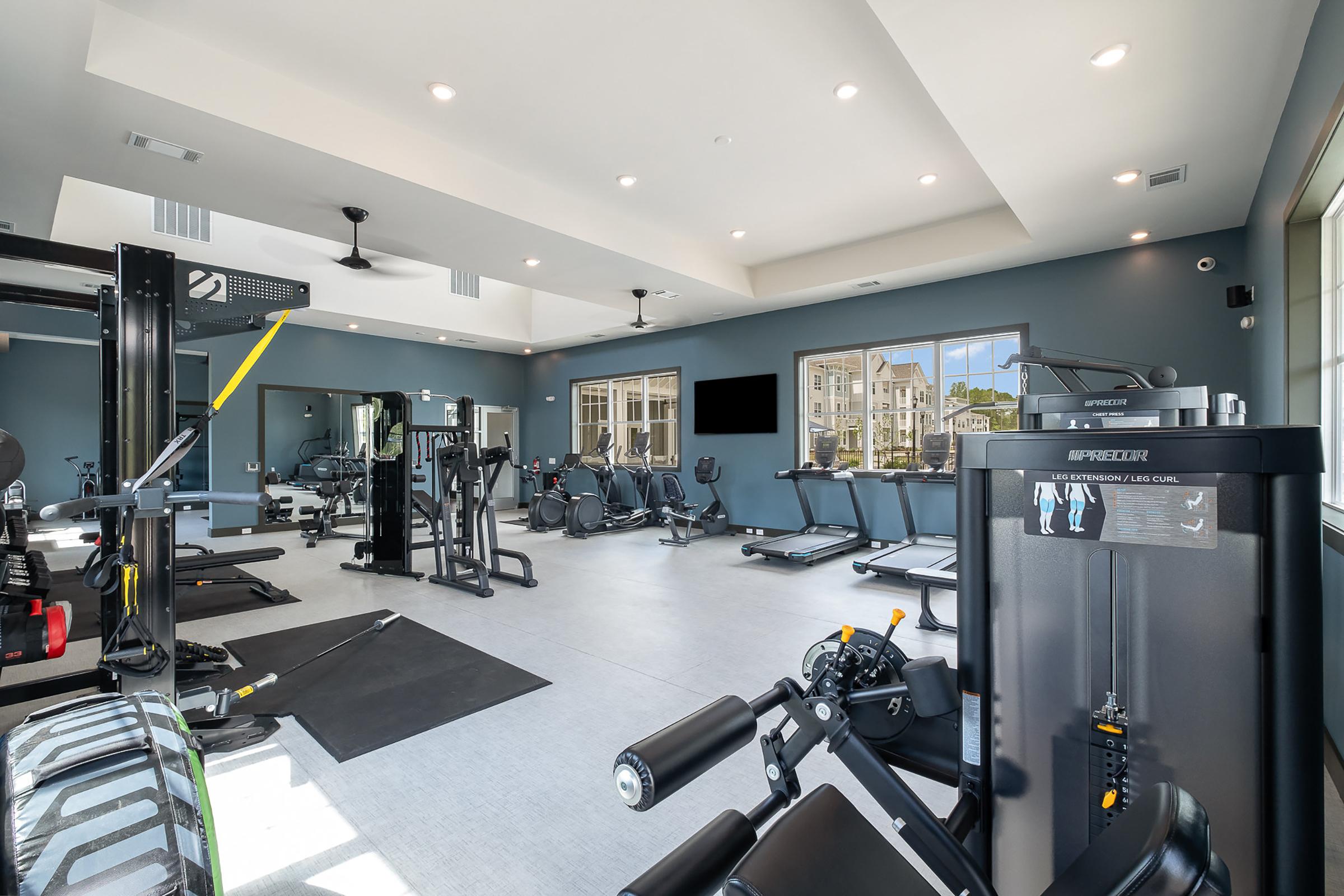 A modern gym interior featuring various workout equipment such as cardio machines, weightlifting stations, and free weights. The spacious area is well-lit with large windows and has a contemporary design, including black and gray color schemes.
