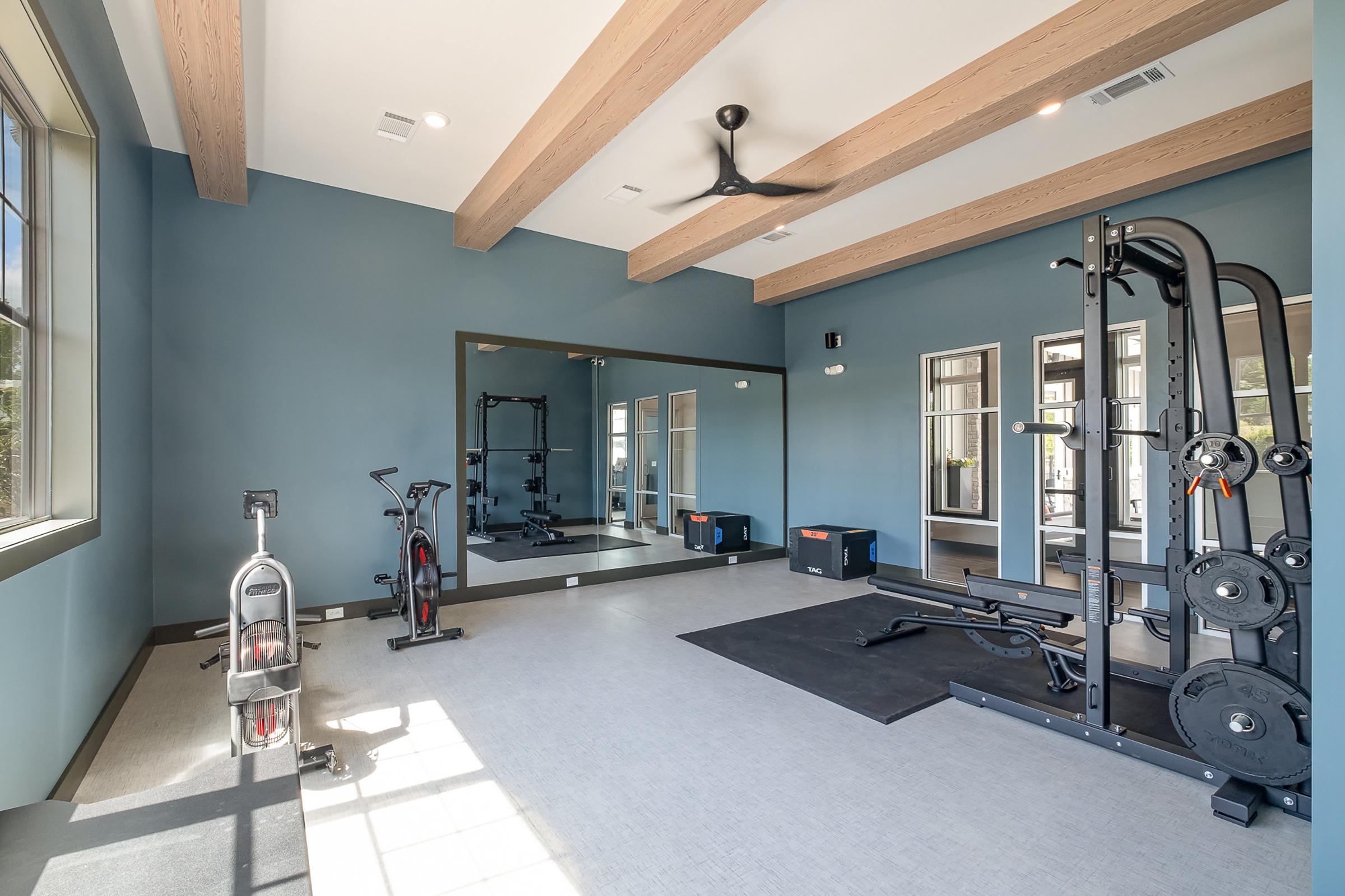 A modern home gym featuring a variety of exercise equipment, including a stationary bike, weight bench, and cable machine. Large windows allow natural light to fill the space, and wooden beams accent the ceiling. A large mirror reflects the workout area, enhancing the sense of openness.