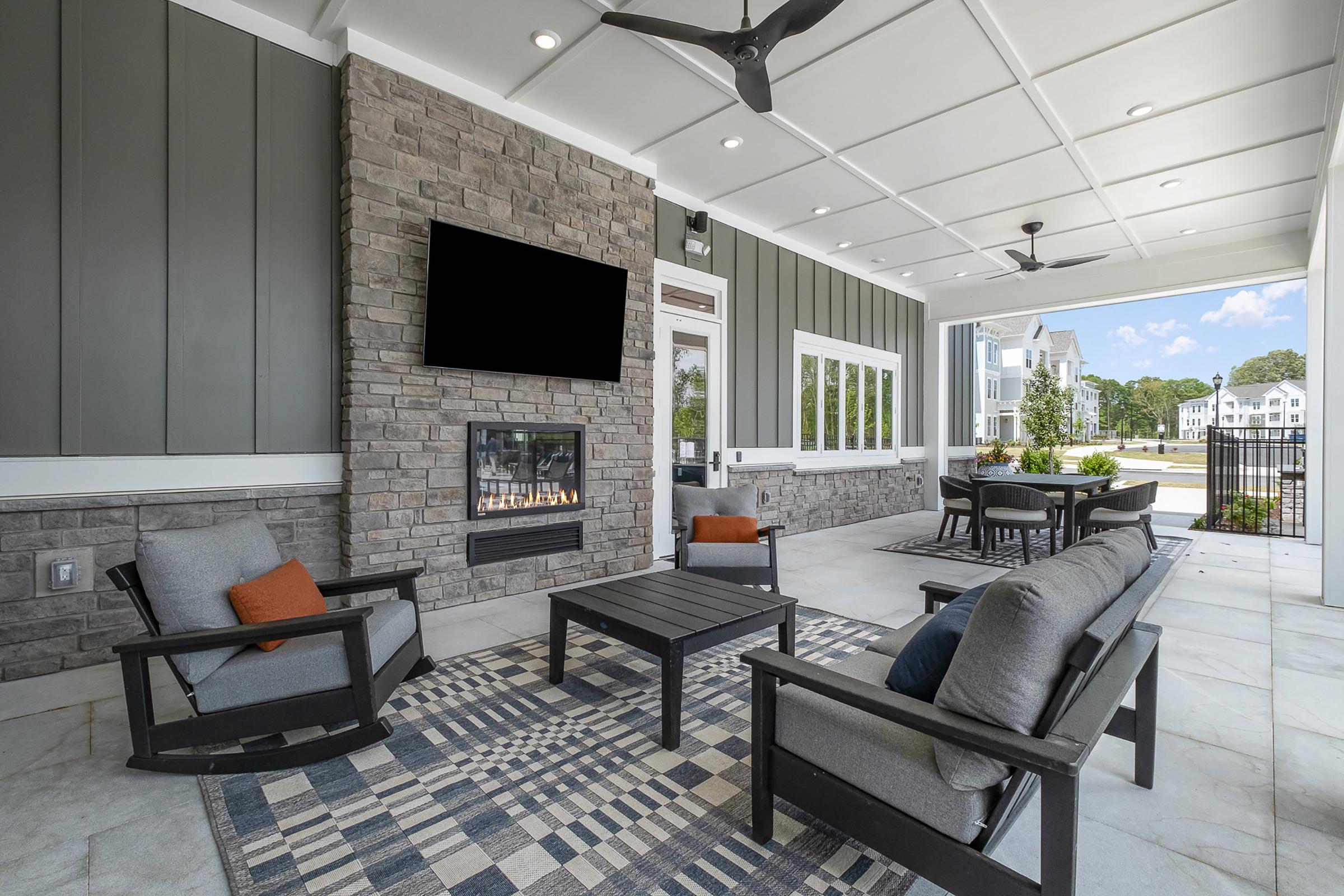 A spacious outdoor lounge area featuring gray seating with orange cushions, a coffee table, and a stone fireplace. A large television is mounted on the wall, and the space is well-lit with natural light coming from large windows, overlooking a pleasant neighborhood setting.