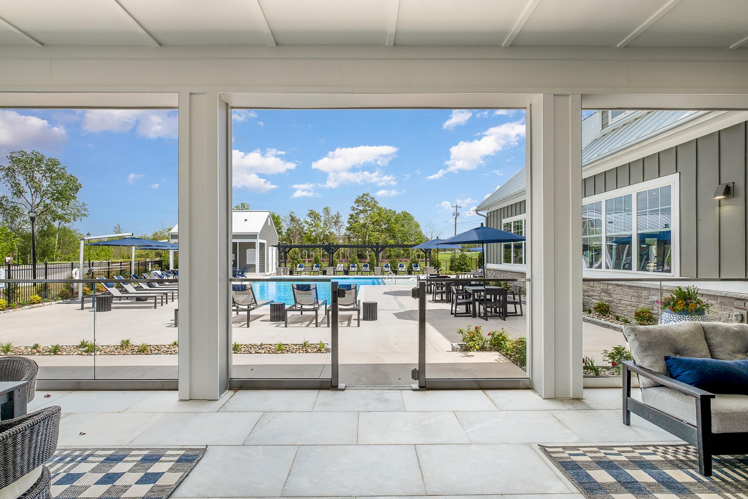 A modern outdoor patio view featuring a swimming pool surrounded by lounge chairs and tables. In the foreground, there are comfortable seating options with pillows. The scene is bright with blue skies and greenery in the background, creating a relaxing atmosphere.