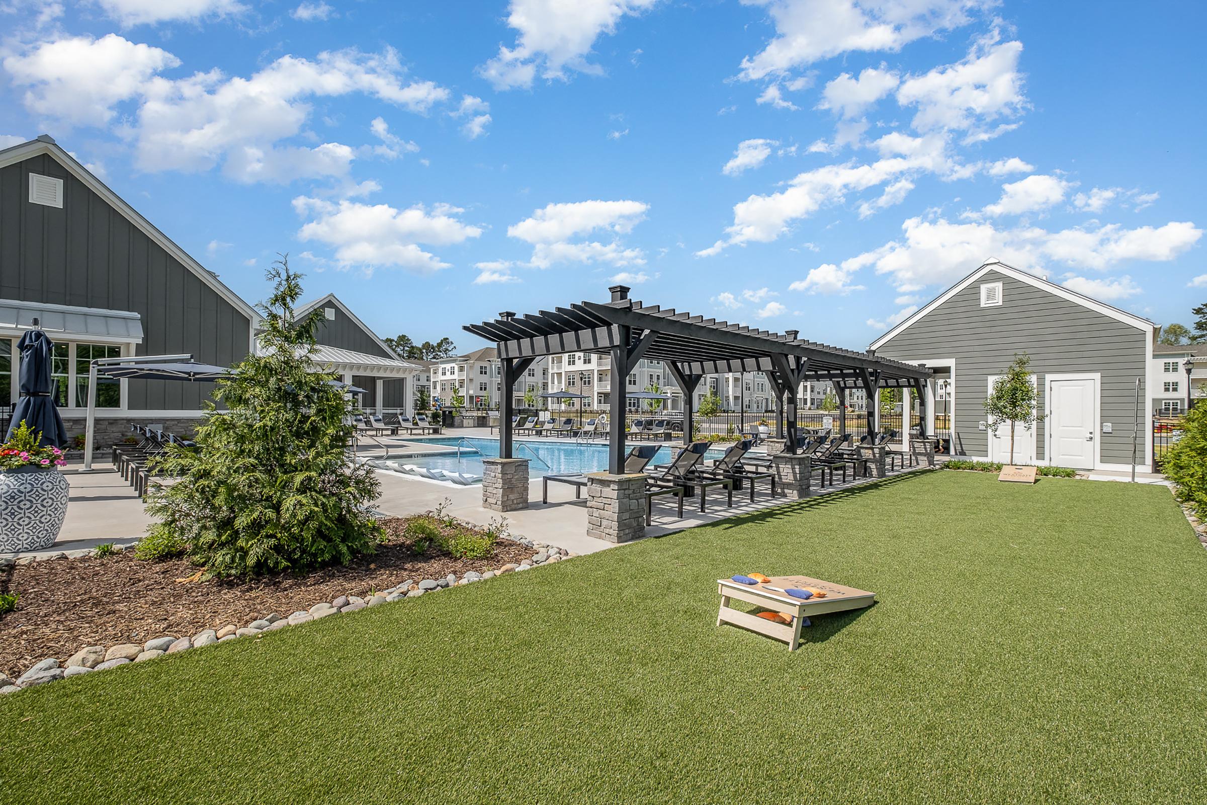 A beautifully landscaped outdoor space featuring a swimming pool surrounded by lounge chairs and shaded seating areas. There are green grass and plants, with a cornhole game set up on the lawn. Elegant residential buildings are visible in the background under a clear blue sky with scattered clouds.