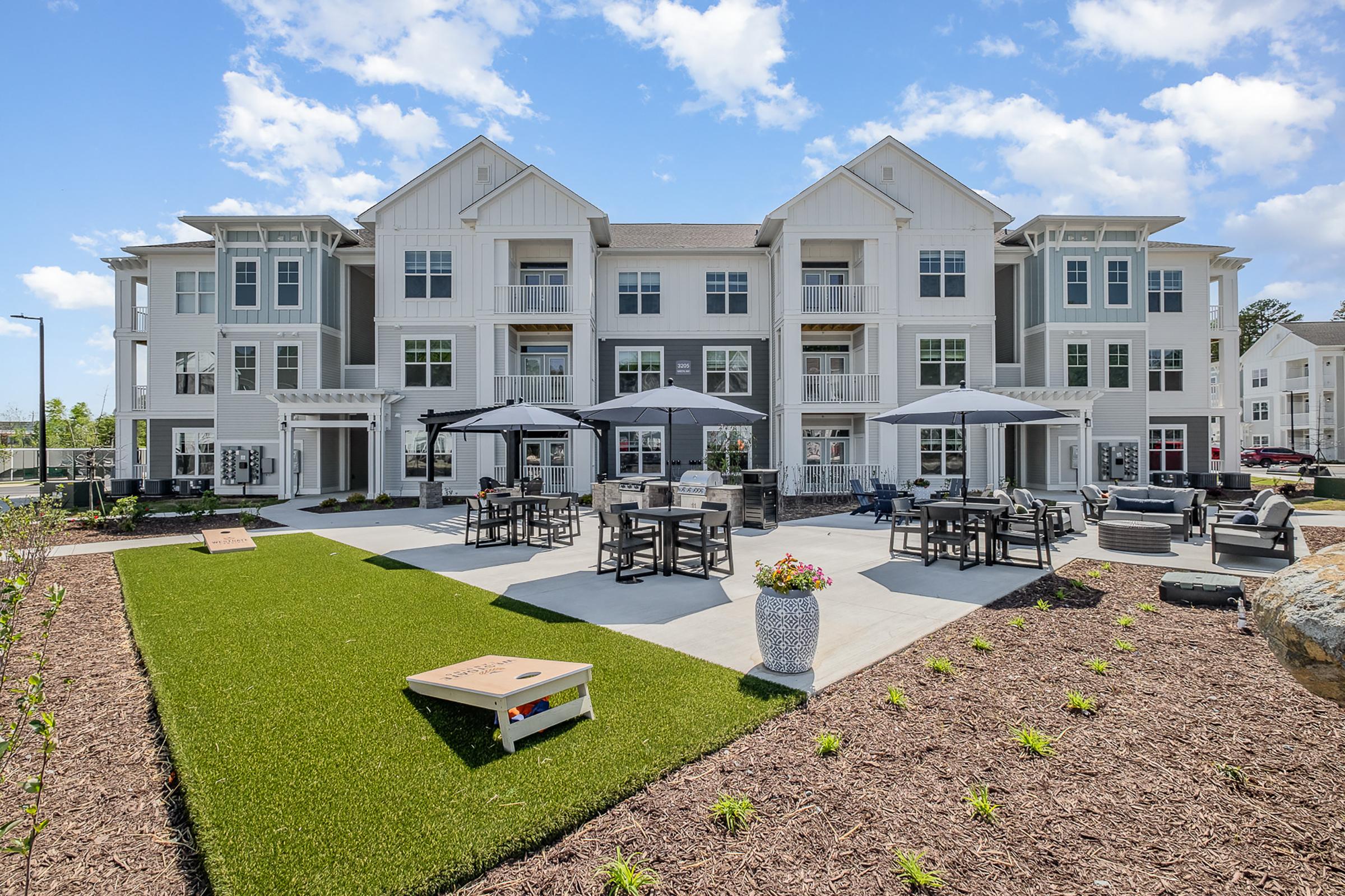 A modern multi-story apartment building with a landscaped outdoor area featuring seating, tables, and umbrellas. The patio is surrounded by green grass and decorative plants, creating a welcoming atmosphere for residents to relax and socialize. Bright blue sky complements the scene.