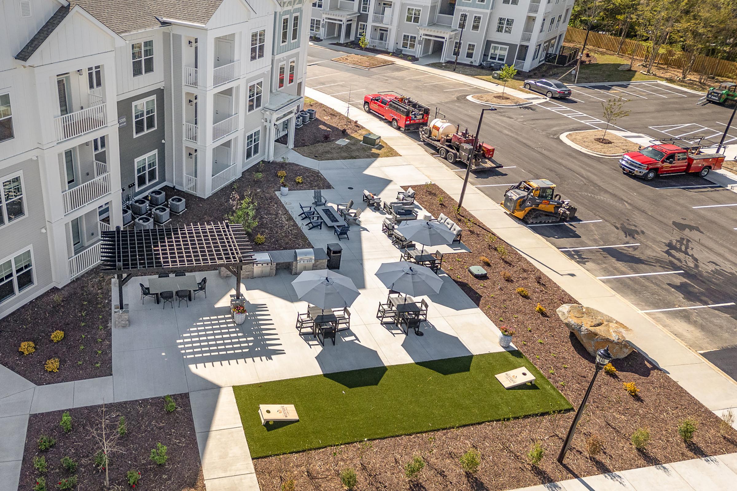 Aerial view of a residential area featuring an outdoor patio with black metal chairs and tables, shaded by large umbrellas. Nearby are freshly landscaped areas with grass and plants. Construction vehicles are parked in the lot, and several buildings can be seen in the background.
