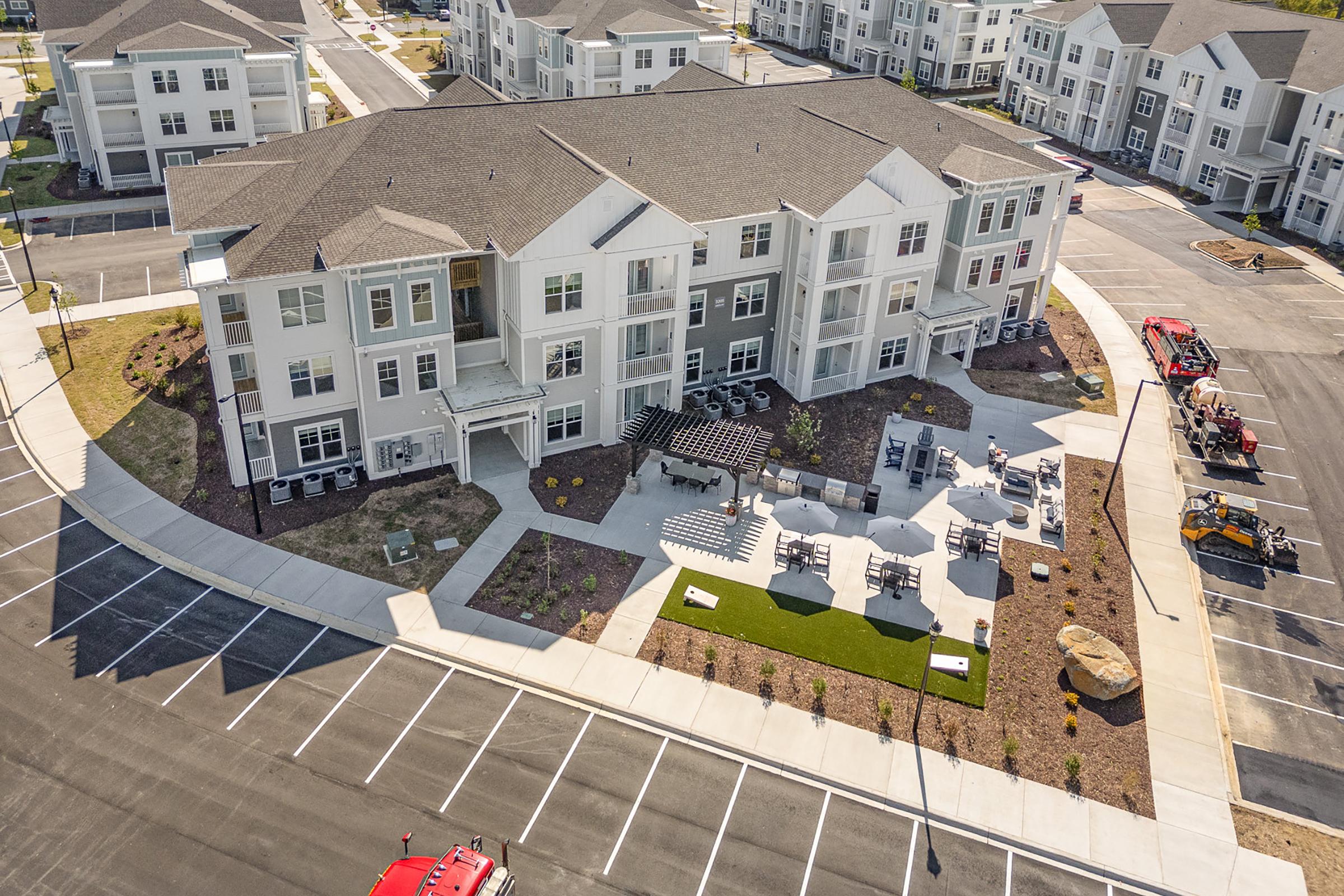 Aerial view of a modern apartment complex featuring a landscaped courtyard with seating areas, outdoor furniture, and stone features. The surrounding area includes paved parking spaces and additional buildings in the background.