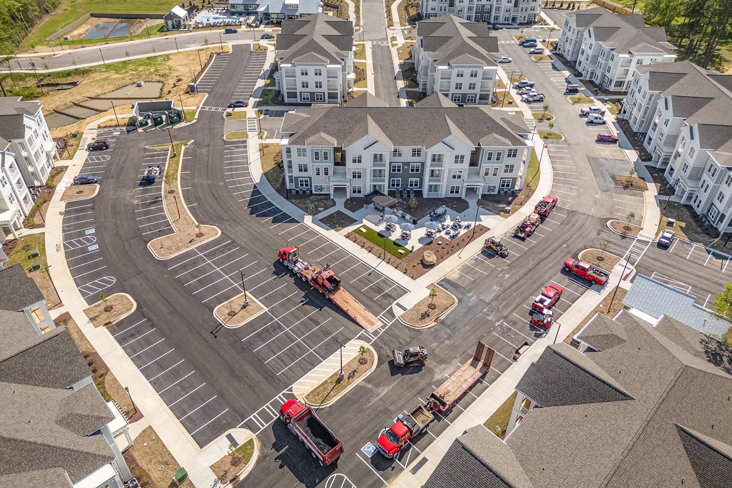 Arial view of a newly developed residential area featuring several multi-story apartment buildings. Numerous vehicles, including trucks, are parked in the newly paved lot, with some landscaping work in progress. The layout includes roads, parking spaces, and green areas, suggesting a planned community environment.
