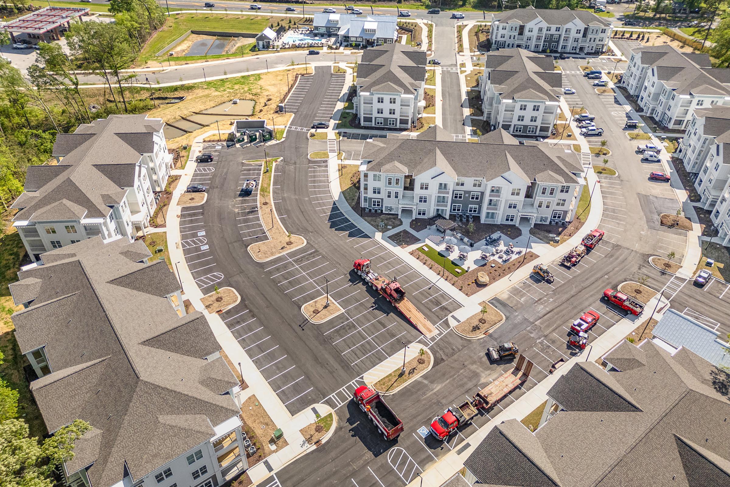 Aerial view of a residential complex under construction. The layout features several multi-story buildings, paved roads, and parking areas. Construction equipment and vehicles are parked nearby, indicating ongoing development. Green landscaping is visible in the surroundings.