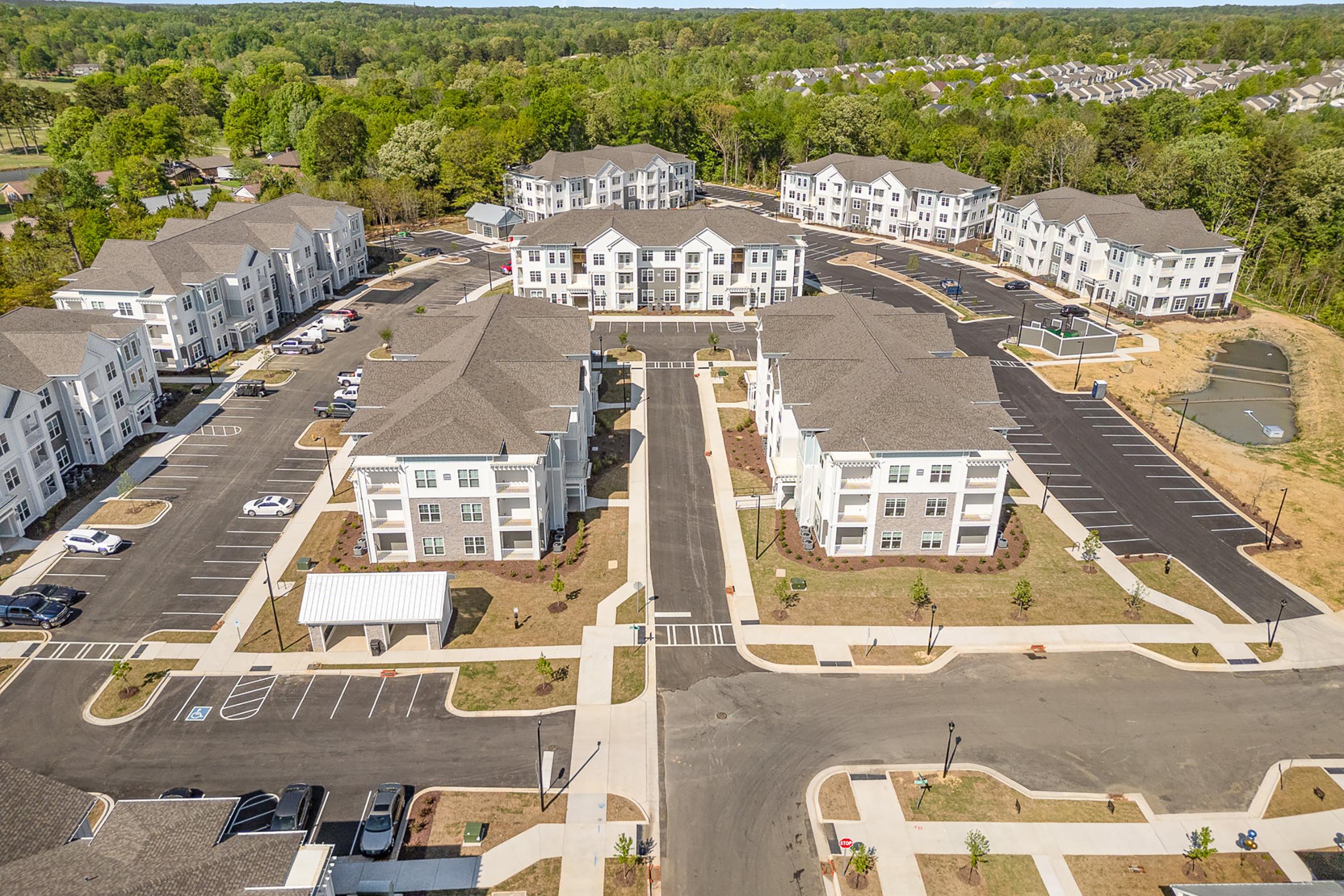 Aerial view of a modern apartment complex featuring multiple buildings with landscaped grounds. The layout includes parking areas, paved walkways, and green spaces, surrounded by trees and natural scenery. The surrounding area shows additional residential buildings in the background.