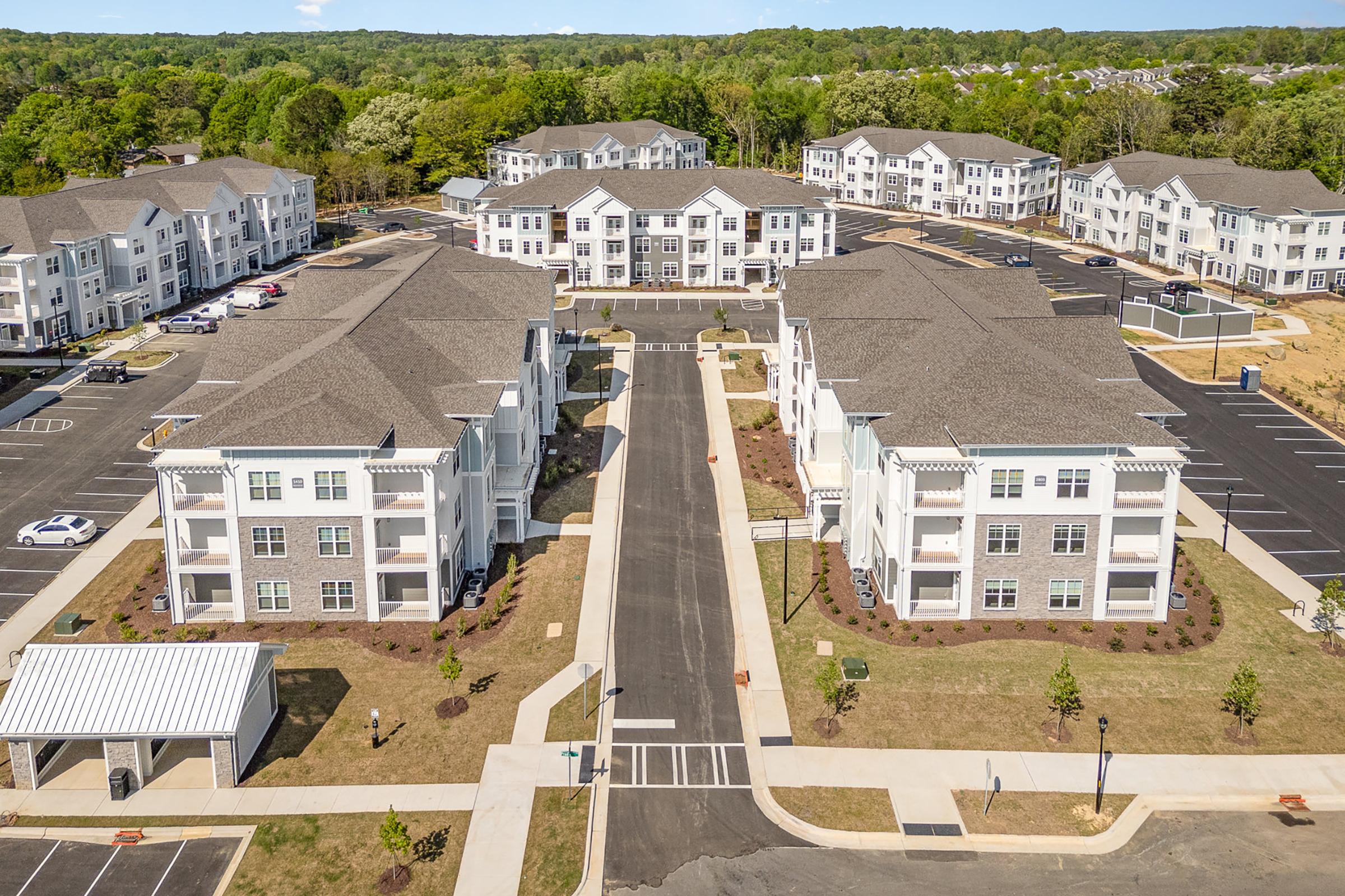 Aerial view of a modern apartment complex featuring several multi-story buildings arranged around a central road. The landscape includes landscaped areas, parking spaces, and surrounding trees, indicating a suburban setting. Clear blue sky with green foliage in the background.