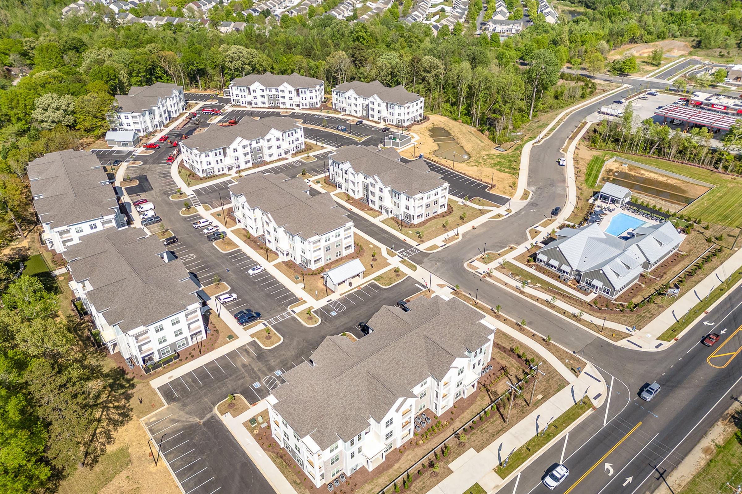 Aerial view of a suburban residential development featuring multiple two- and three-story apartment buildings arranged around landscaped parking areas. The community is surrounded by greenery and includes well-maintained roadways and sidewalks.