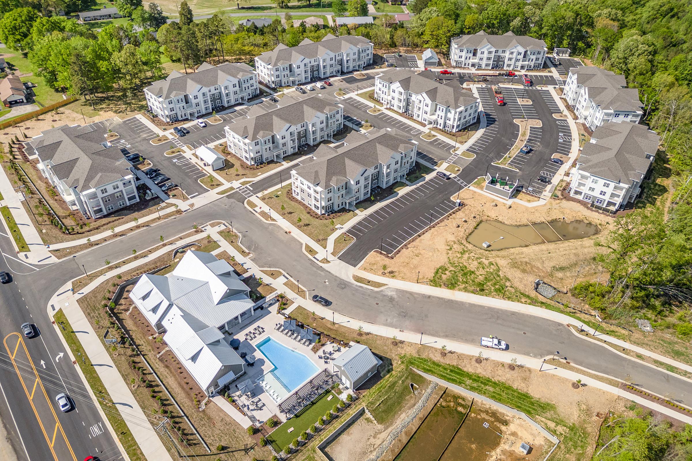 Aerial view of a modern residential complex featuring several multi-story apartment buildings, a central community area with a swimming pool, and landscaped green spaces. Surrounding parking and roadways are visible, along with areas of undeveloped land. Trees and vegetation are present in the background.