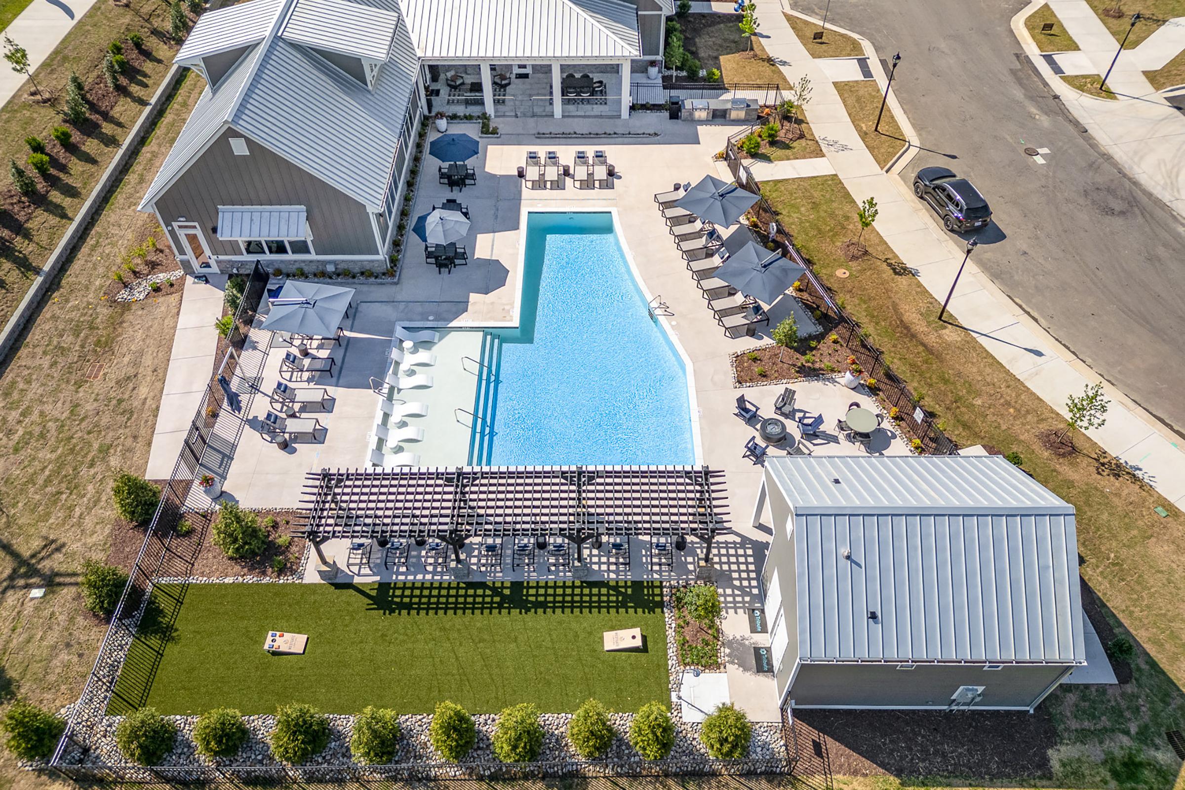 Aerial view of a community pool area featuring a large blue swimming pool surrounded by lounge chairs and umbrellas. Adjacent is a grassy area with seating and cornhole games. There are two buildings nearby, one with a gray roof and the other with a lighter shade, set within a landscaped environment.