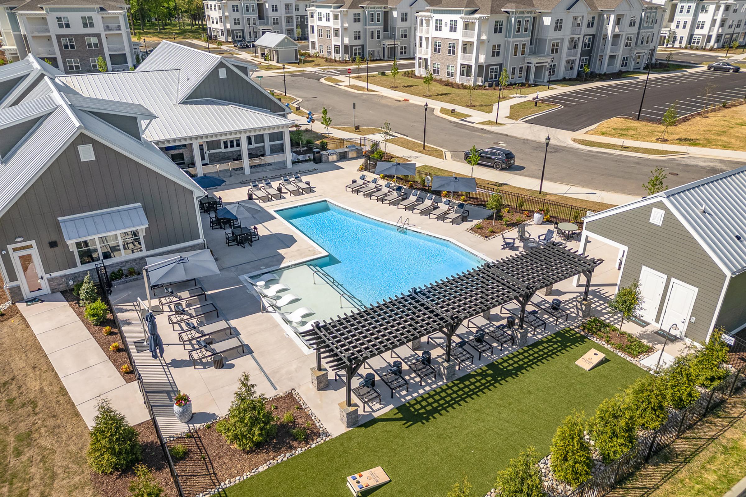 Aerial view of a modern apartment complex featuring a large swimming pool surrounded by lounge chairs. Nearby, there are shaded seating areas with pergolas and well-manicured lawns. The setting includes several residential buildings in the background, showcasing a community atmosphere.
