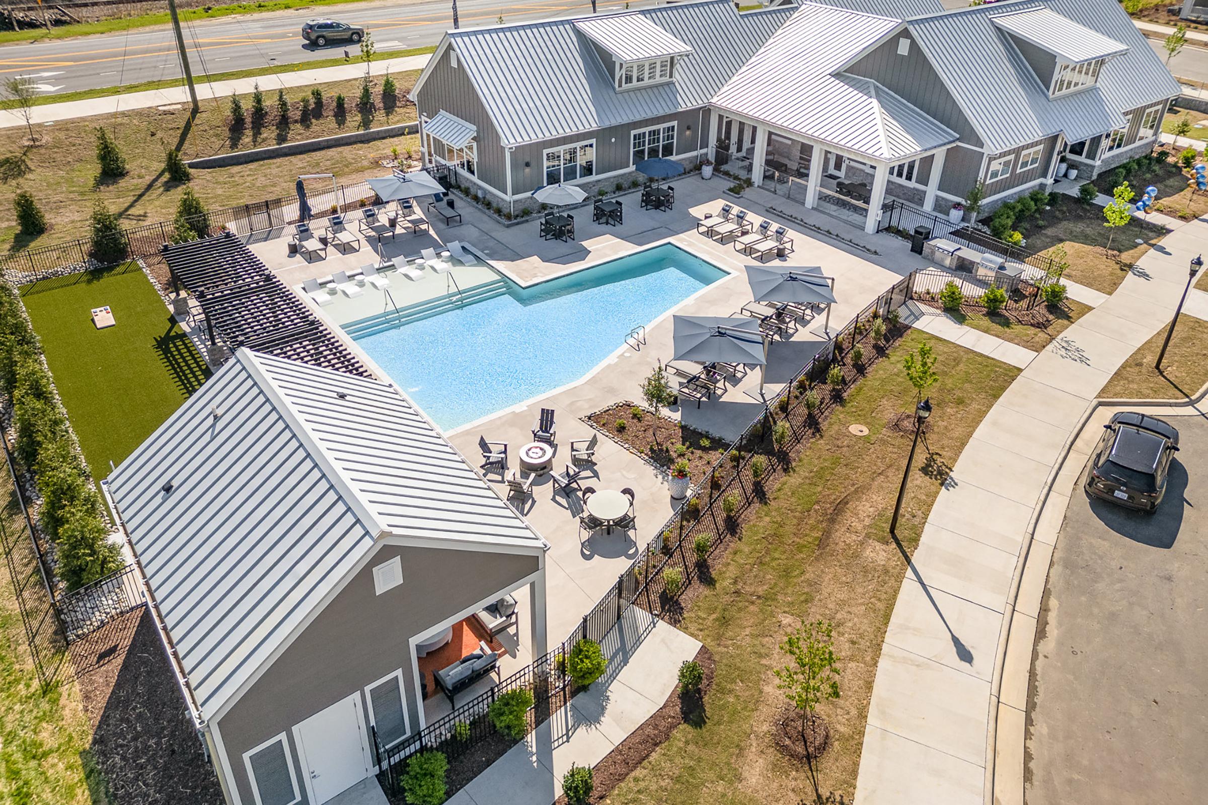 Aerial view of a residential community featuring a swimming pool surrounded by lounge chairs and umbrellas. Nearby, there are two buildings with modern architecture and a grassy area, all set in a well-maintained landscape. The scene is sunny and inviting, showcasing amenities for leisure and relaxation.