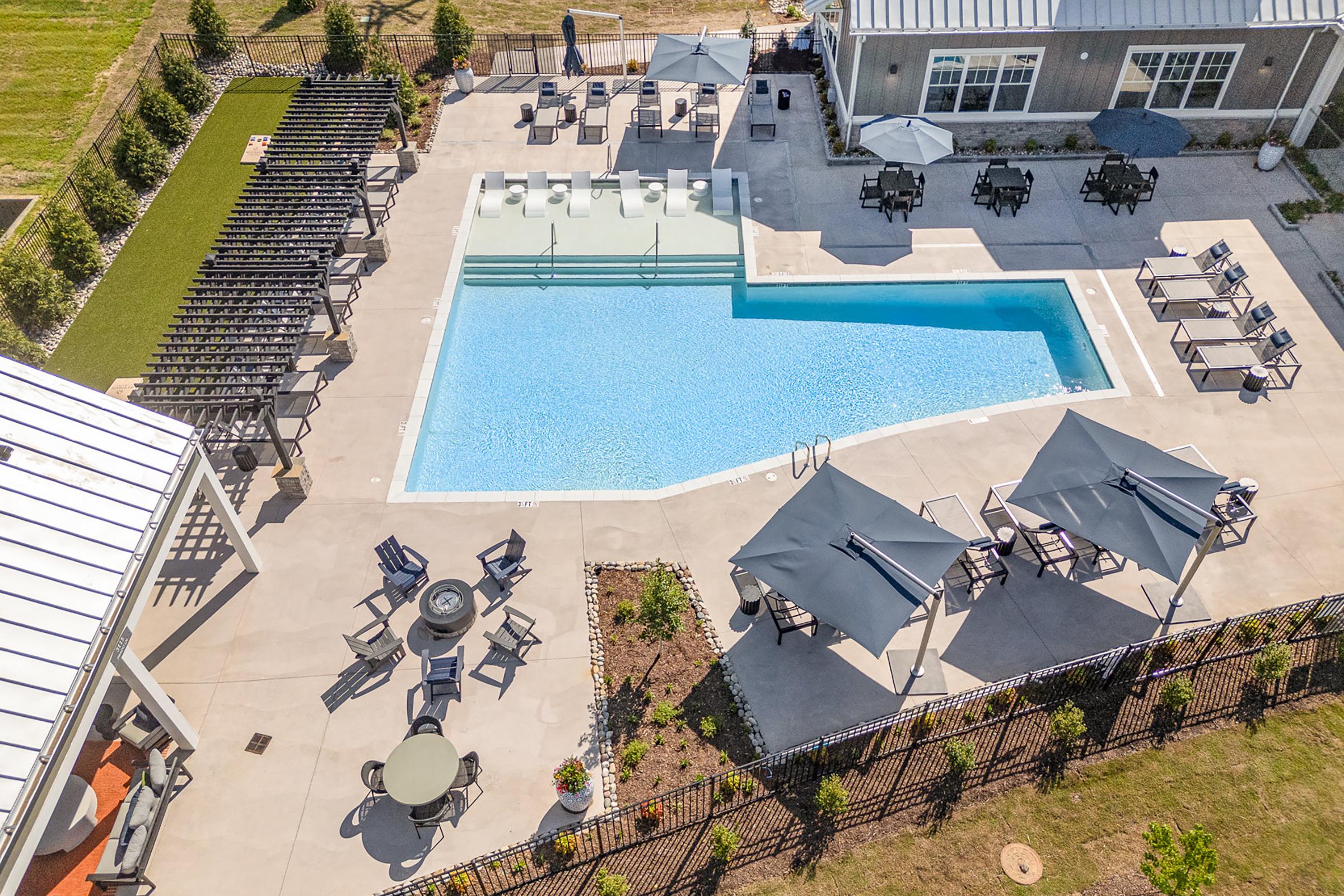 Aerial view of a modern pool area featuring a clear blue swimming pool surrounded by lounge chairs and shaded tables with umbrellas. The area includes neatly arranged landscaping with flowers and grassy patches, alongside a building visible in the background.