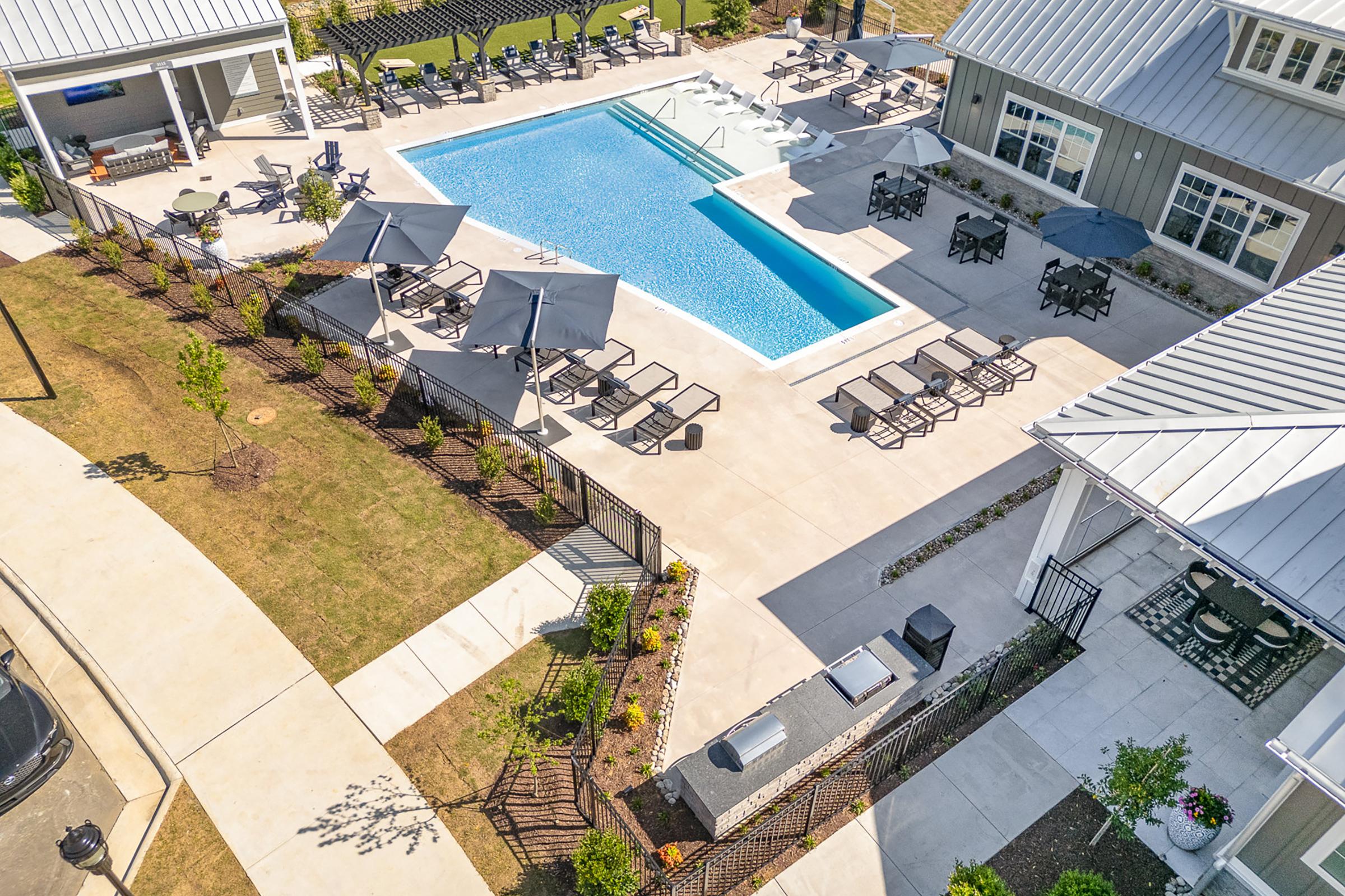 Aerial view of a pool area featuring a large, rectangular swimming pool surrounded by lounge chairs and umbrellas. There are well-maintained gardens and pathways connecting to a nearby building. The area is designed for relaxation and leisure, with a grill station visible for outdoor cooking.