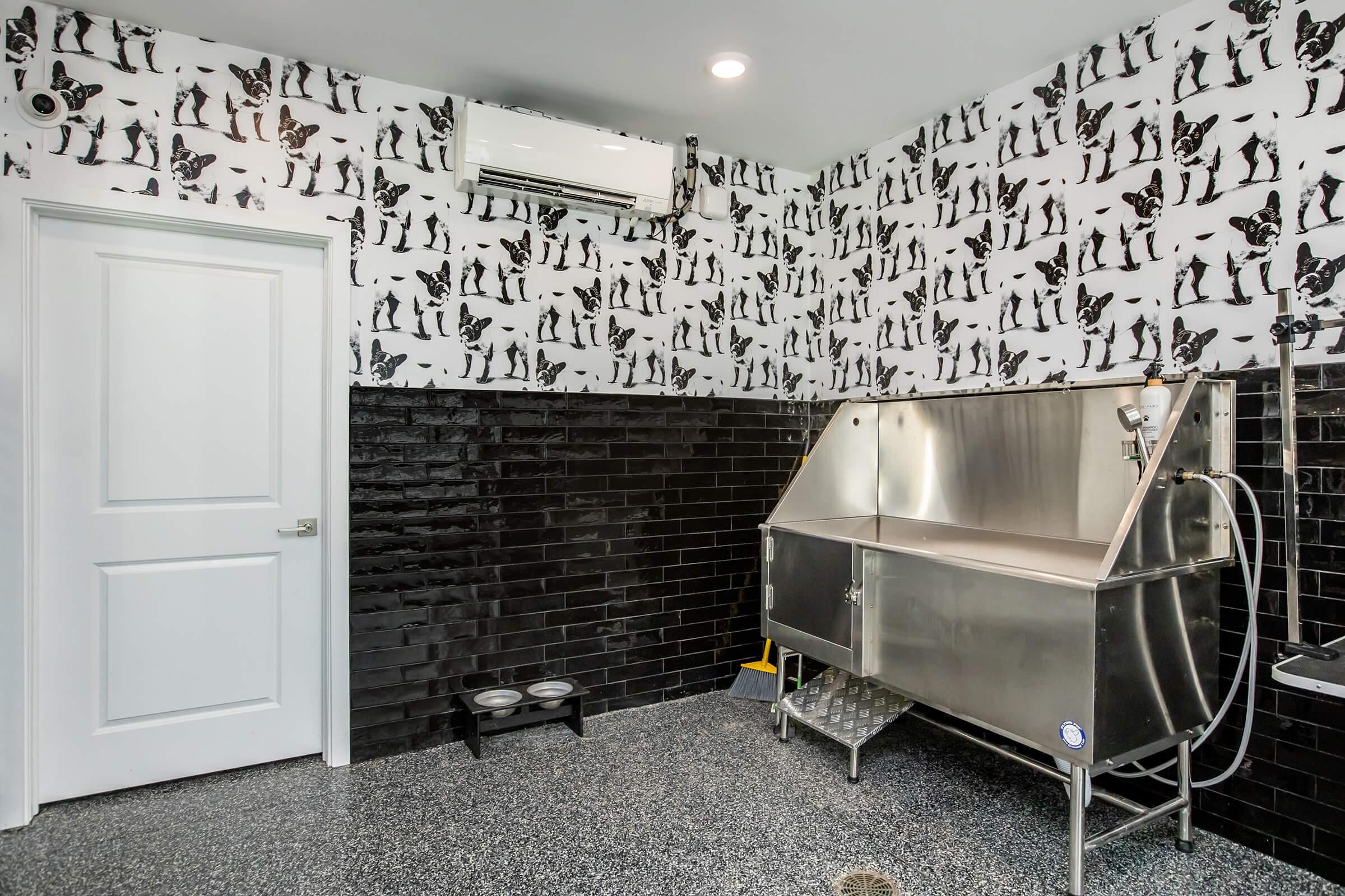 Interior of a pet grooming room featuring black and white cat and dog patterned wallpaper. Includes a stainless steel grooming tub, a wall-mounted air conditioner, a white door, and a floor with a drainage system. There are food and water bowls on the floor.