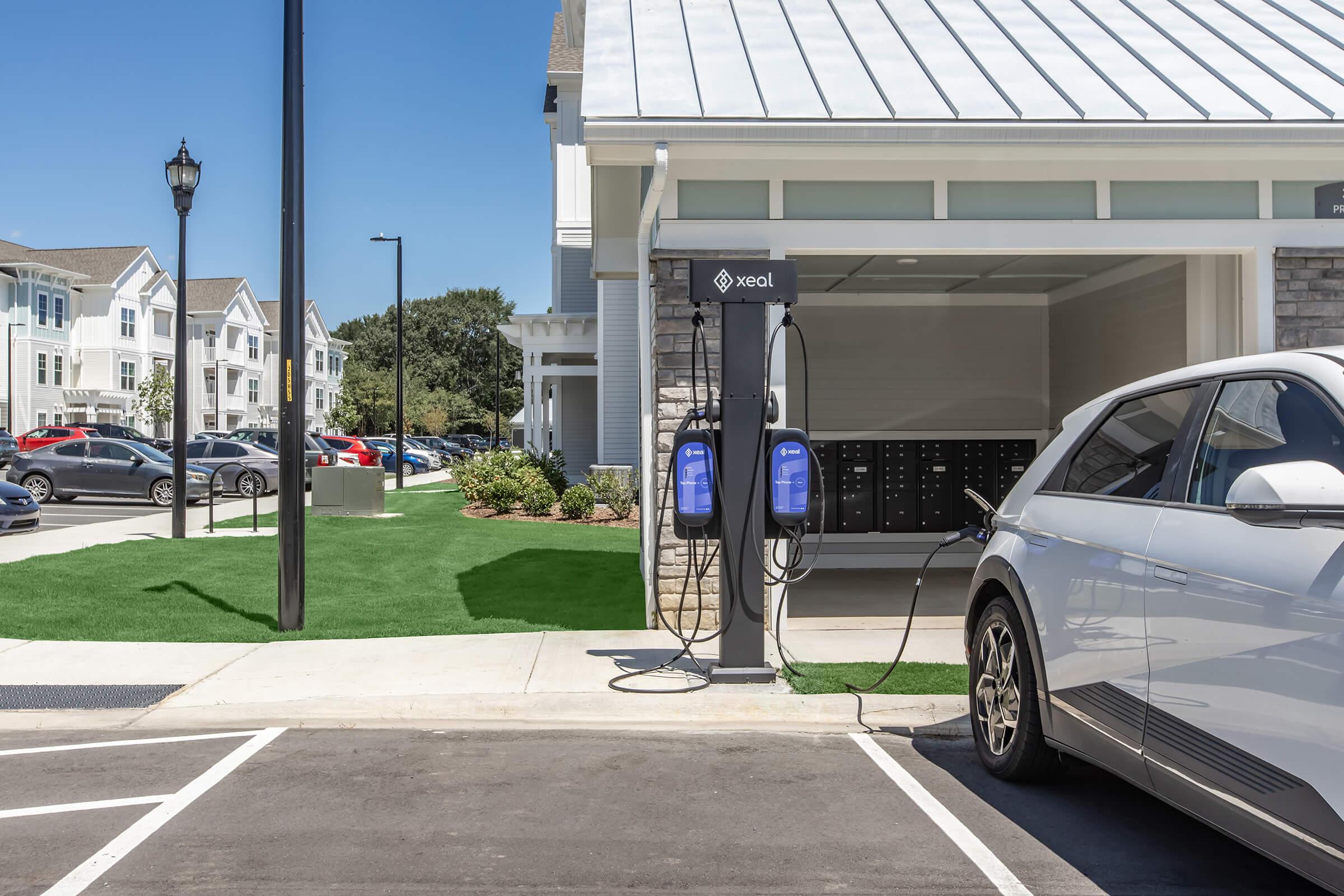 Electric vehicle charging station beside a modern apartment building. A white car is plugged into the charger, which features two charging ports. In the background, several cars are parked, and there is green grass and landscaping around the area. The sky is clear and blue.