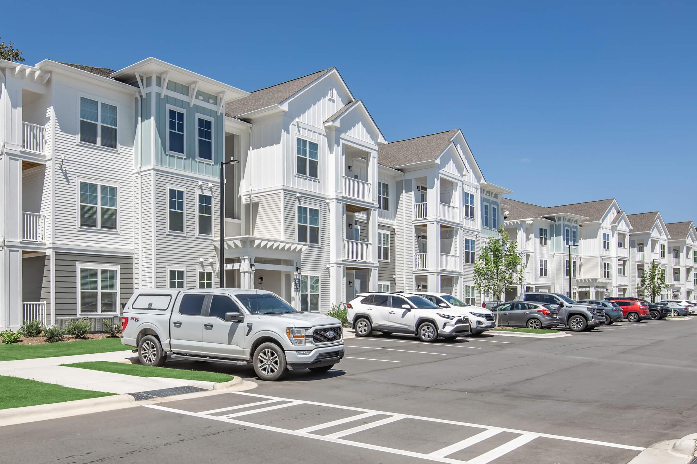 A modern apartment complex with multiple buildings featuring white siding and balconies. The foreground shows a parking lot with various vehicles, including a gray truck and a white SUV. The setting is sunny with clear blue skies and manicured grass.