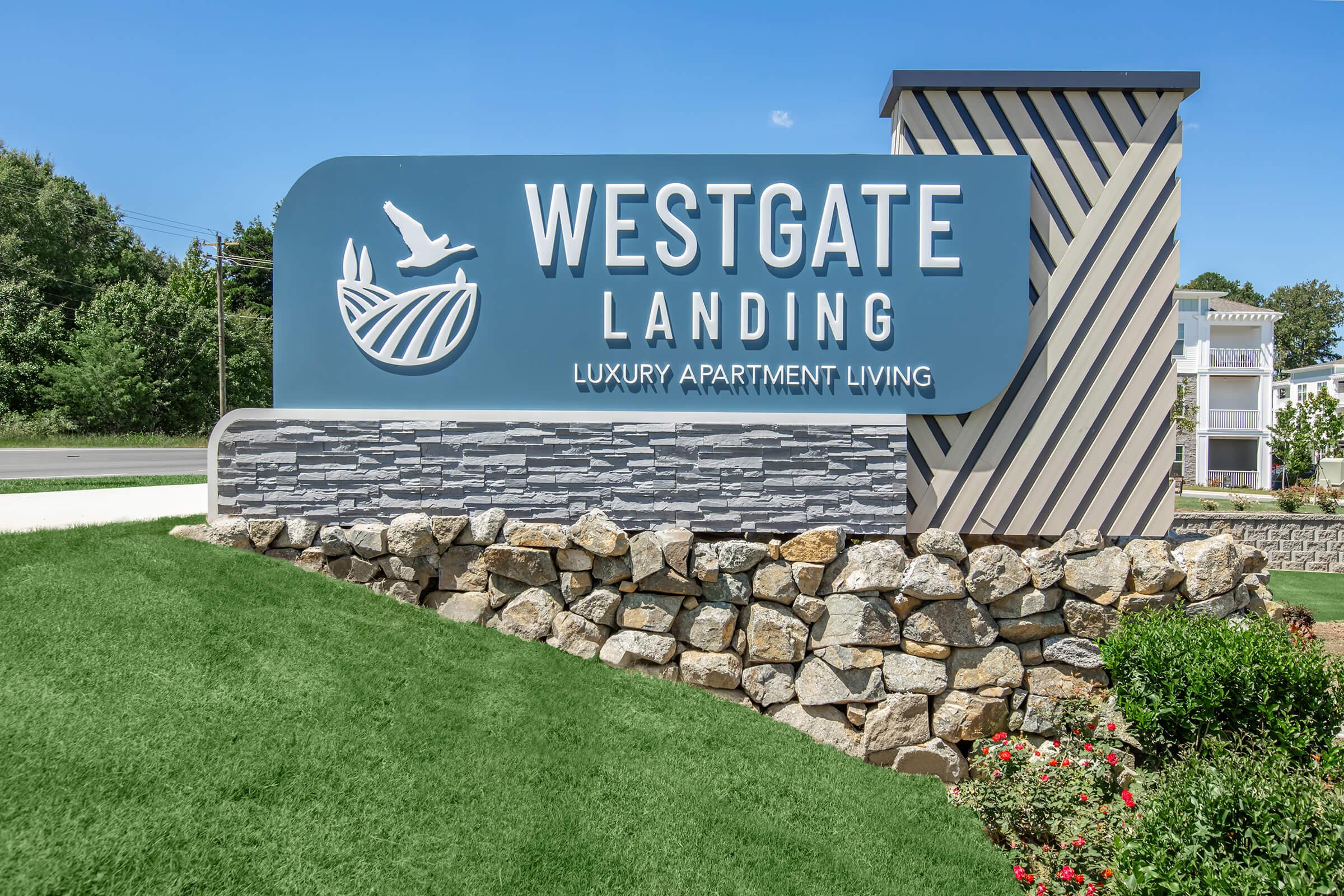 Sign for Westgate Landing featuring the text "Westgate Landing" and "Luxury Apartment Living," set against a backdrop of a stone wall and landscaped grass. The design includes a stylized image of a bird and greenery, situated in a bright, sunny outdoor setting.