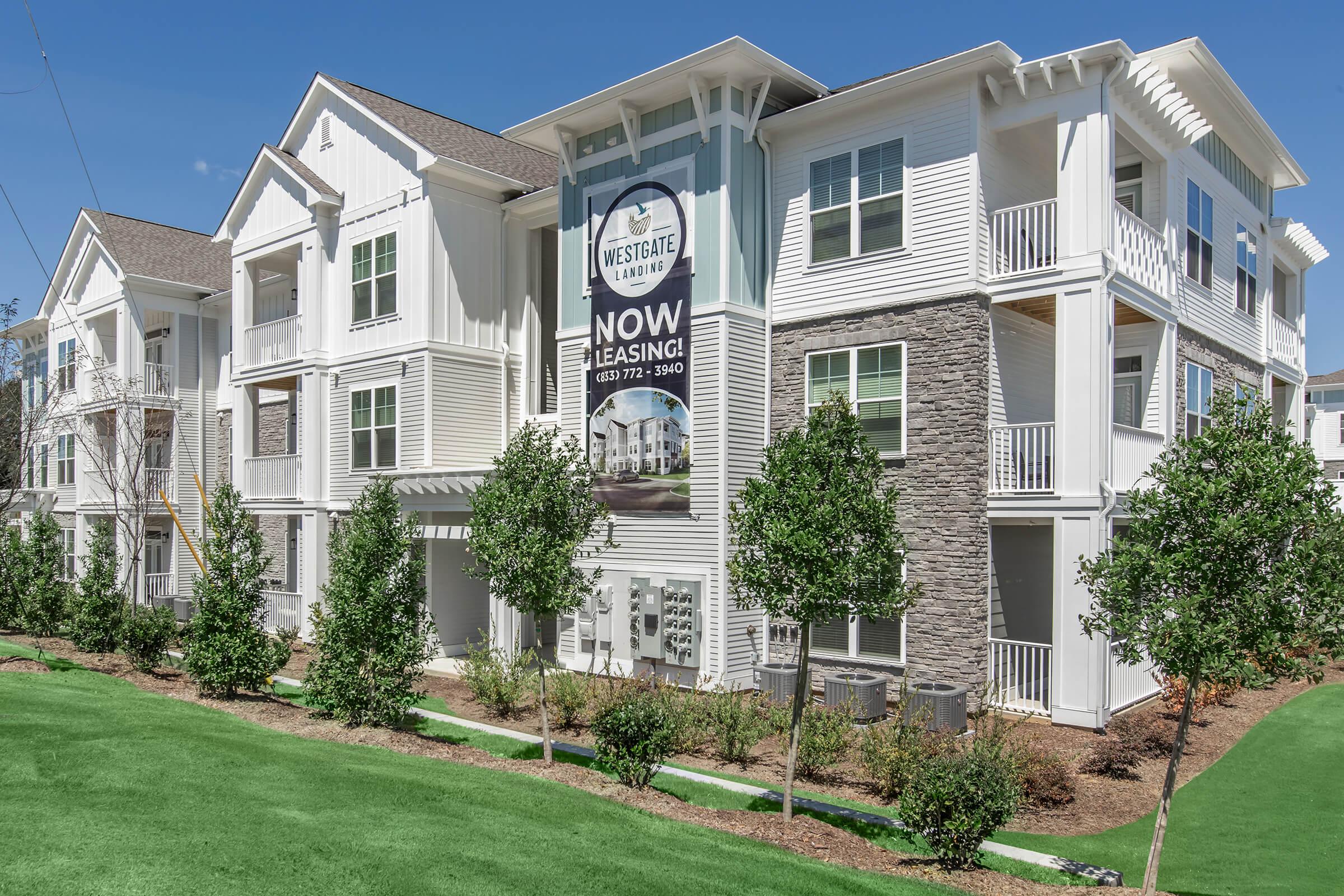 Facade of a modern apartment building with a leasing banner displaying "Now Leasing." The structure features a combination of white siding and stone accents, surrounded by neatly trimmed greenery and landscaping. Clear blue sky is visible above, adding to the bright appearance of the property.