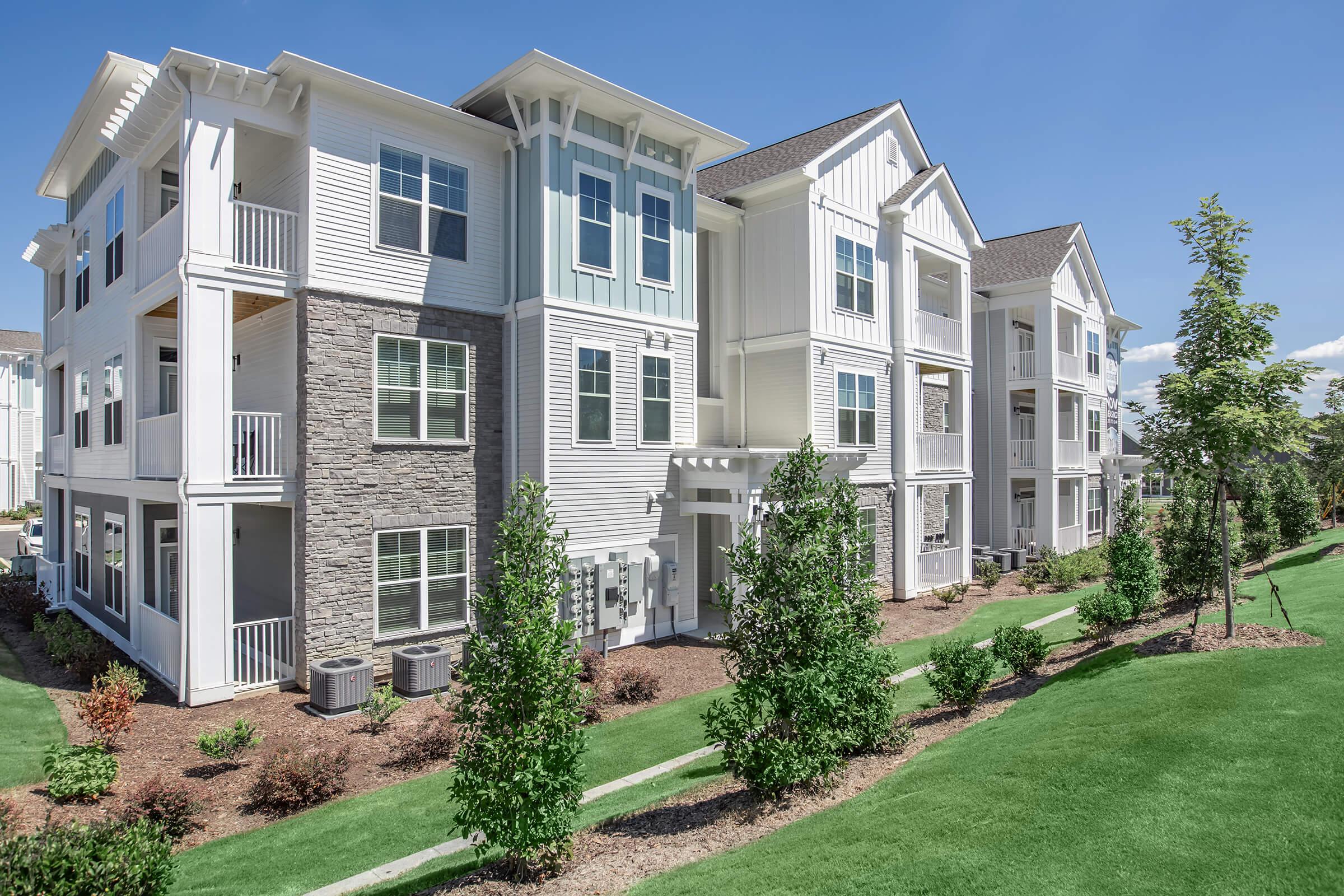 A modern multi-story apartment building featuring a mix of white and stone exteriors with balconies. Lush green landscaping surrounds the building, including neatly trimmed bushes and grass. The sky is clear blue, suggesting a bright and sunny day.