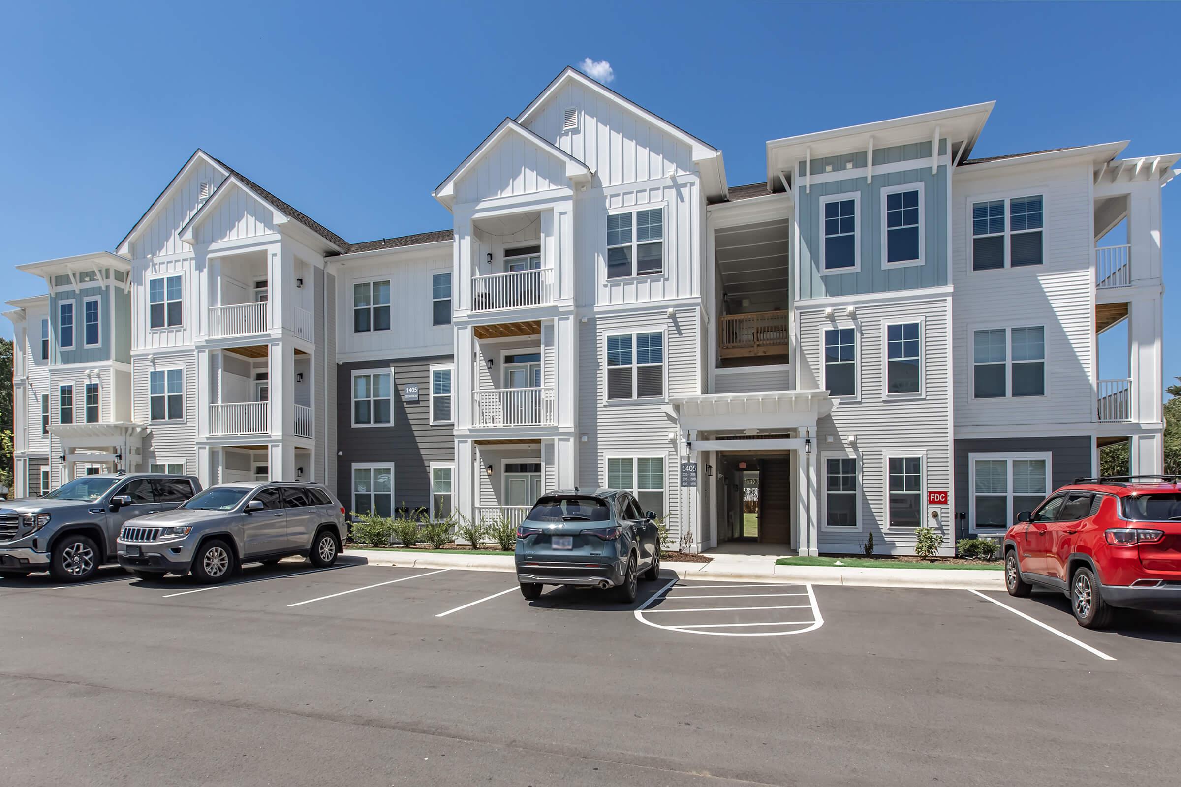 A modern apartment building with a three-story design featuring white and gray siding. The building has multiple balconies and large windows. A parking lot in front has several parked cars. The sky is clear and blue, providing a bright and inviting atmosphere around the property.