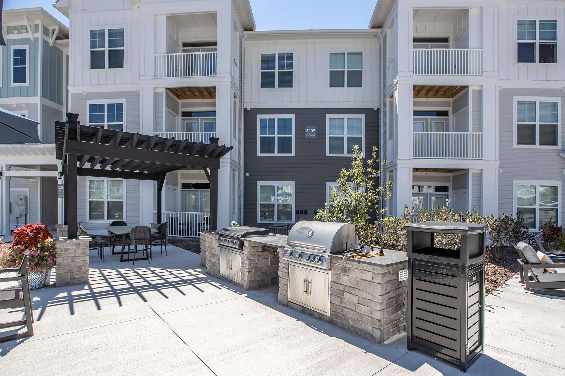 A modern outdoor grilling area featuring two built-in grills and a stone counter, surrounded by a paved patio. There are metal tables and chairs under a pergola nearby, with landscaped greenery and an apartment building in the background. The scene is bright and welcoming, ideal for outdoor gatherings.