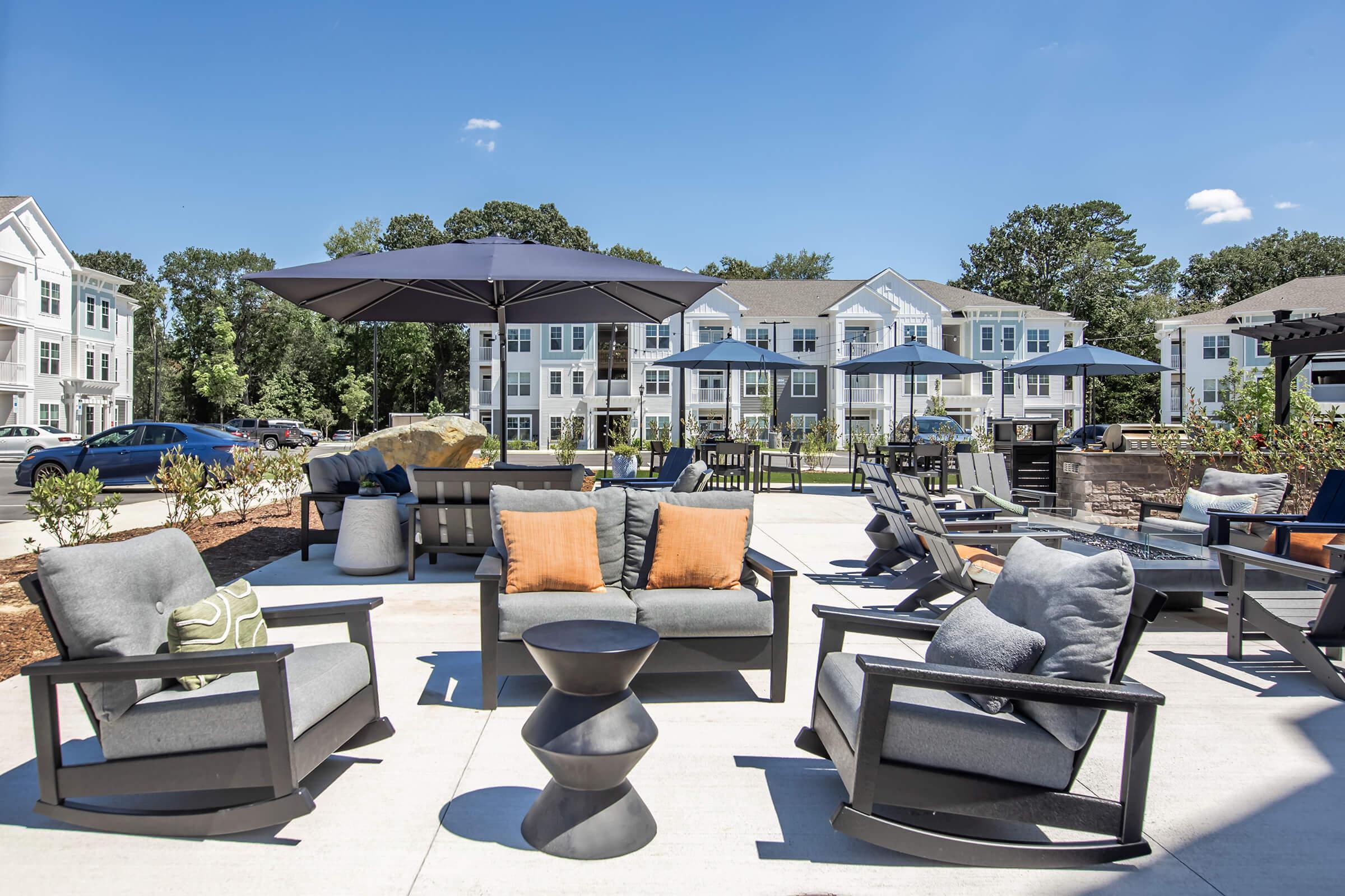 A modern outdoor lounge area featuring stylish gray and orange furniture, including lounge chairs and a small side table. Large umbrellas provide shade over the seating. In the background, there are contemporary apartment buildings surrounded by greenery under a clear blue sky.