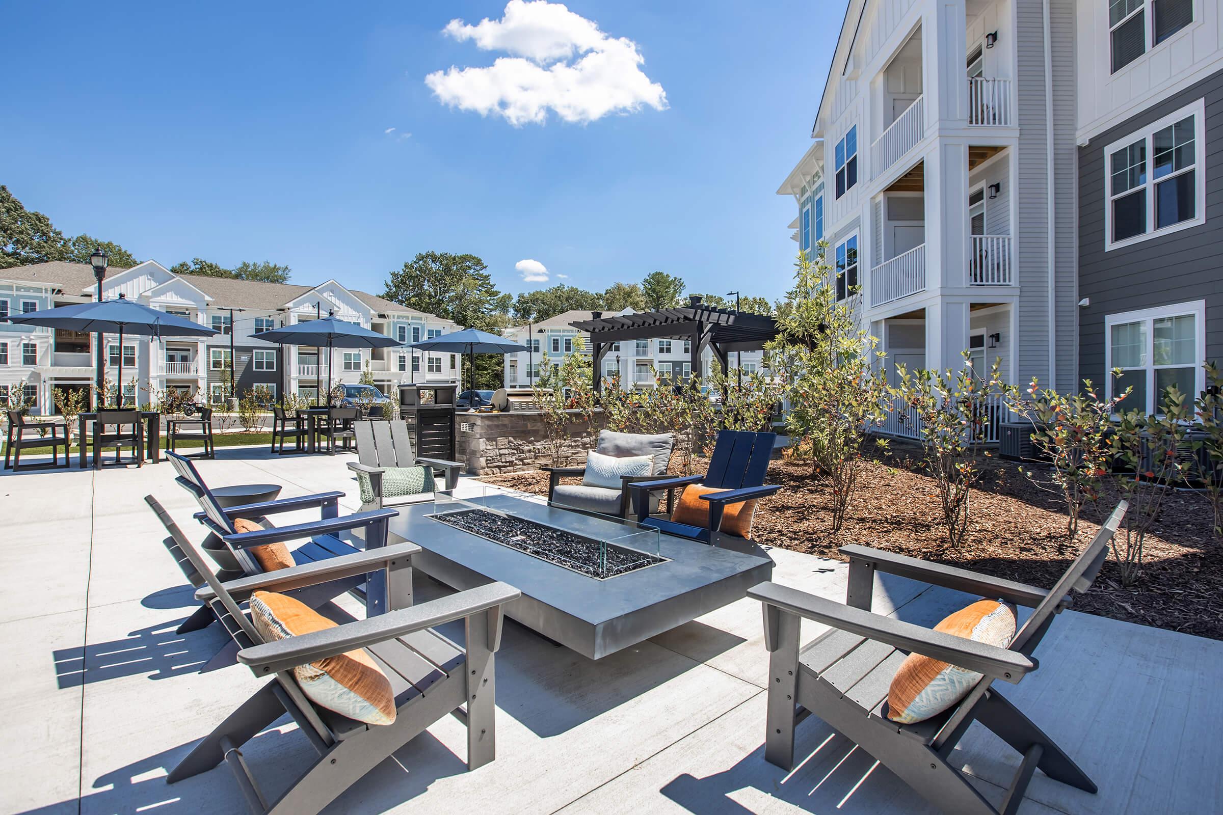 A modern outdoor space featuring a fire pit surrounded by comfortable seating. Patio furniture includes chairs and tables with umbrellas. In the background, multiple multi-story residential buildings and landscaped greenery provide a welcoming atmosphere. Bright blue sky with a few clouds is visible.