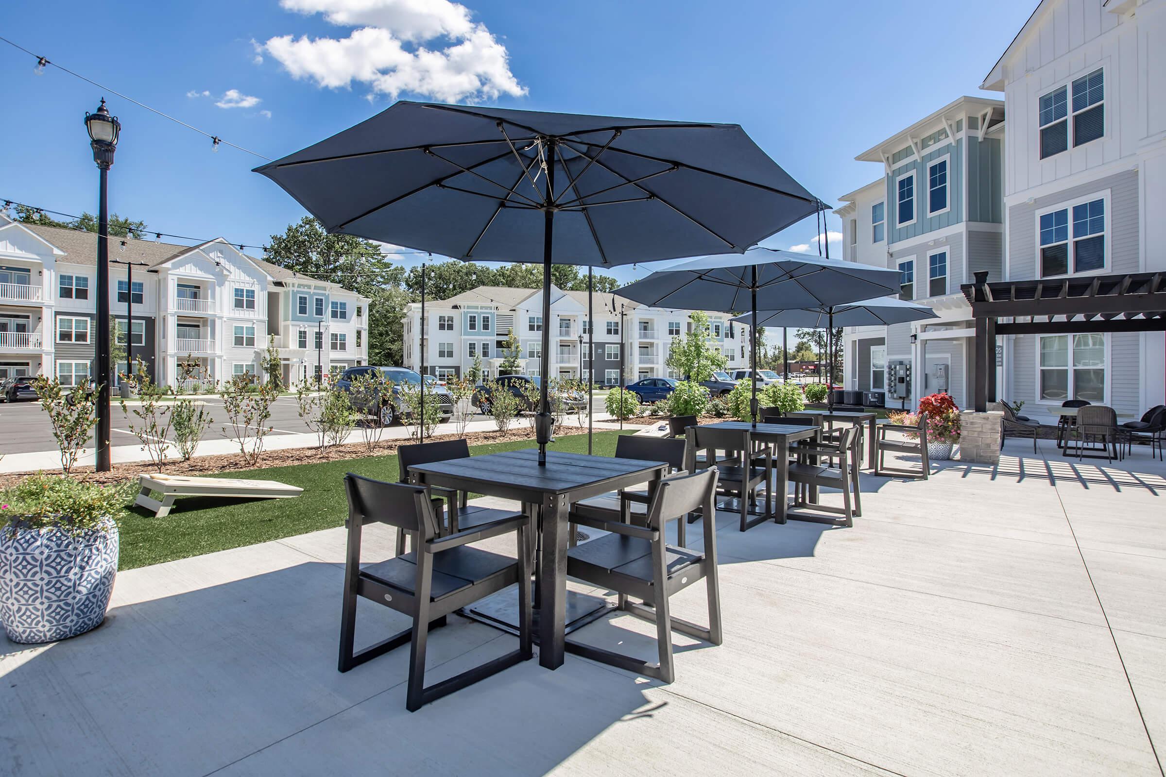 Outdoor seating area with black umbrellas over tables, surrounded by greenery and flowers. Modern apartment buildings are in the background under a clear blue sky, creating a welcoming and sunny atmosphere.