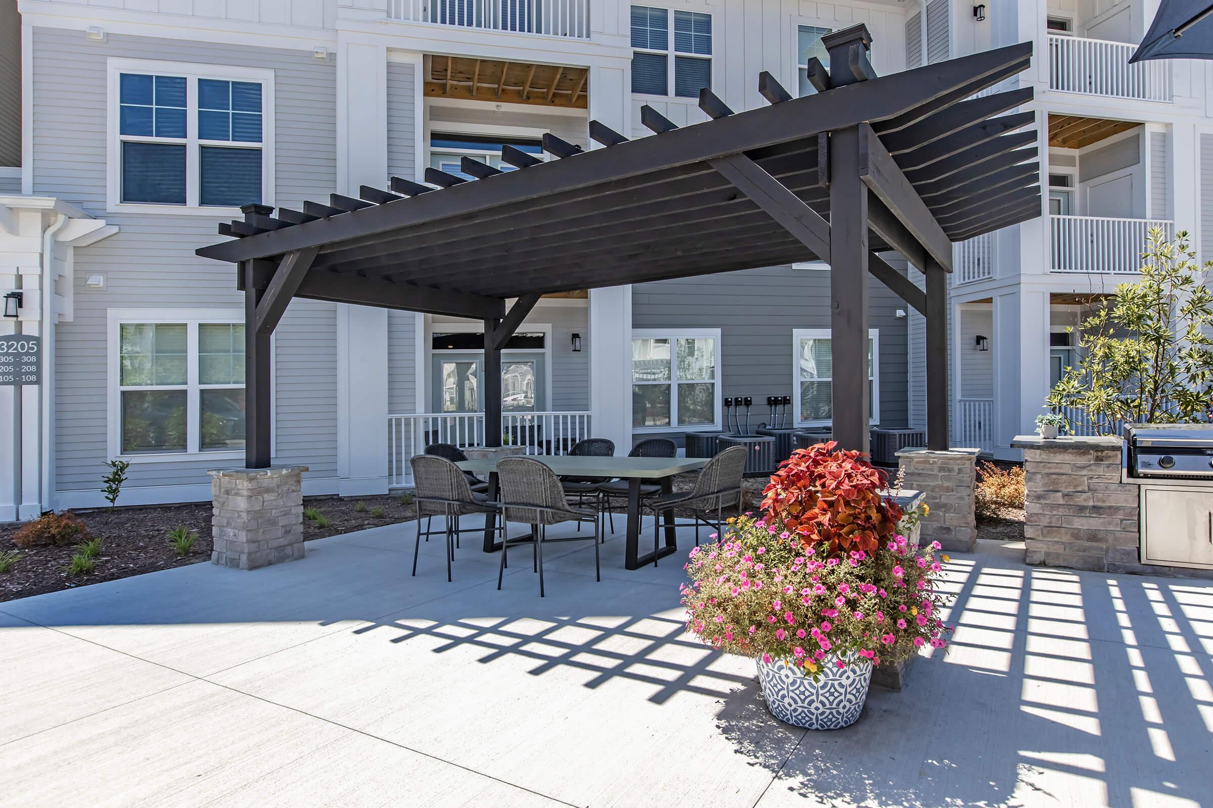 An outdoor dining area featuring a large dark wooden pergola, surrounded by comfortable chairs and a table. A decorative planter filled with colorful flowers is positioned near a stone barbecue grill. The background includes a modern apartment building with large windows and balconies.