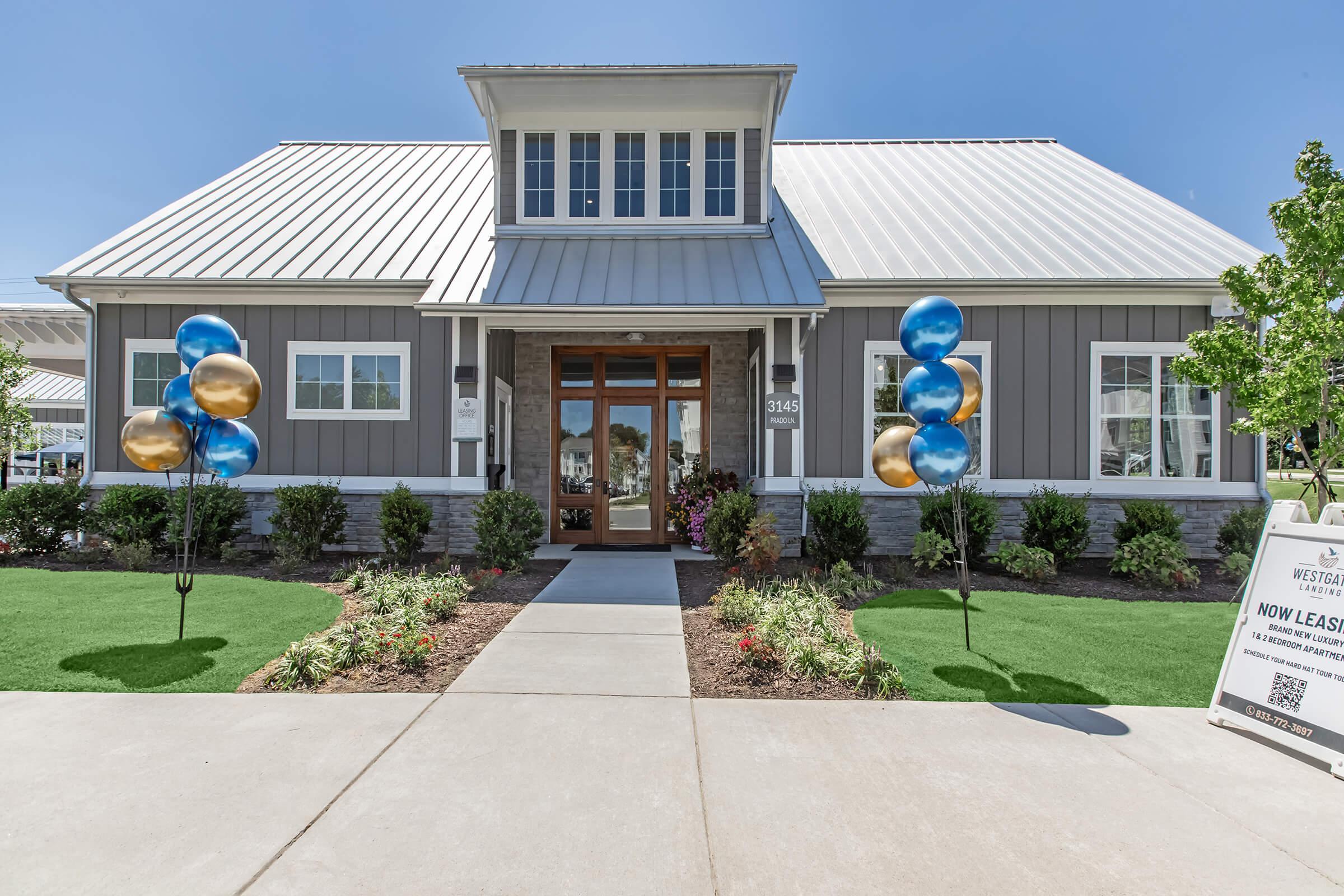 A modern building with a gray exterior and a metal roof, featuring large windows and a welcoming entrance. Flanking the pathway are colorful balloons in blue and gold. A sign indicates that the property is now leasing. Lush greenery and flower beds enhance the inviting atmosphere.