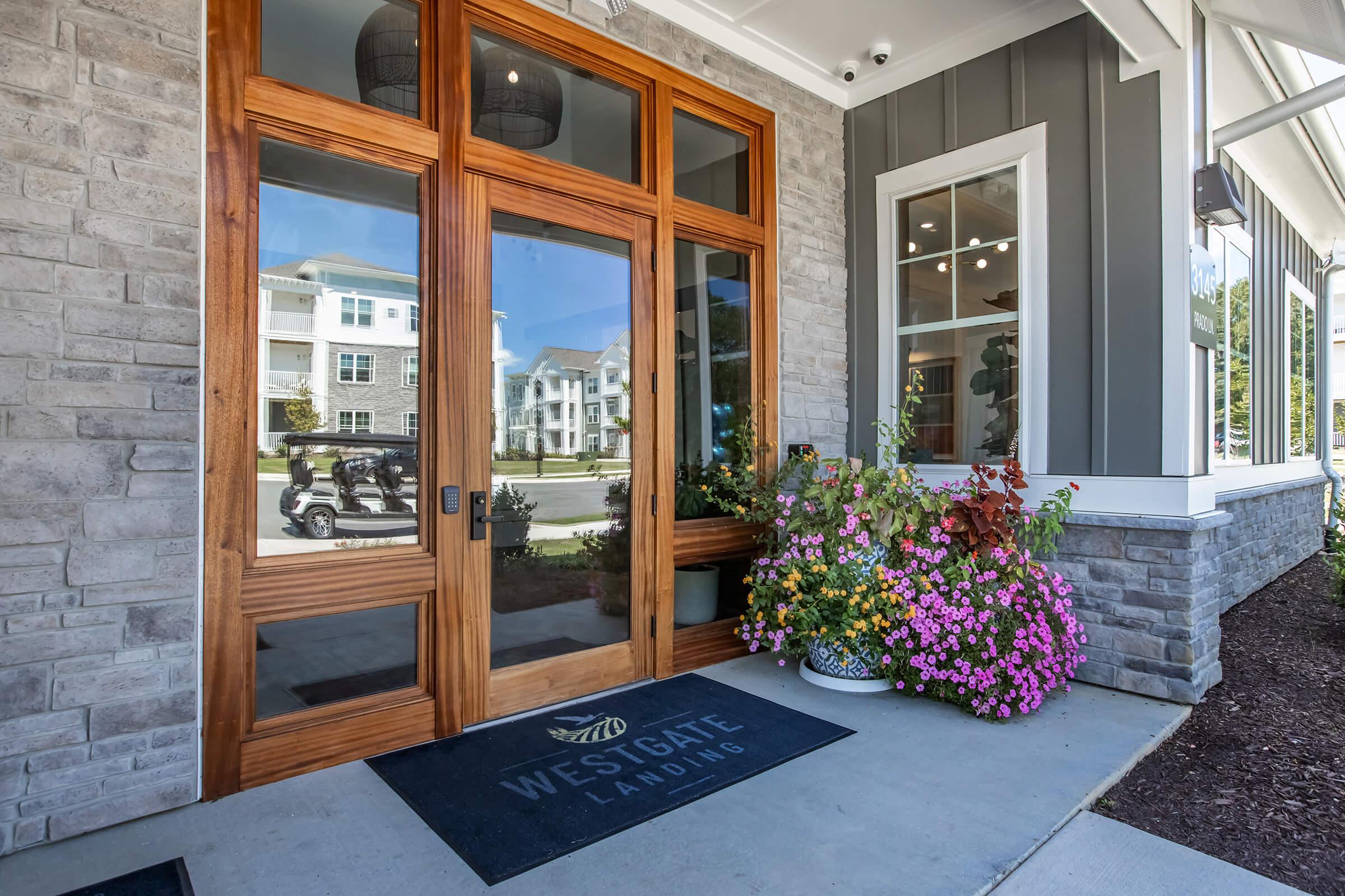 Entrance to Westgate Landing featuring large wooden double doors, a stone facade, and a welcoming flower arrangement beside the entrance. A doormat is visible with the Westgate Landing logo, enhancing the inviting atmosphere.