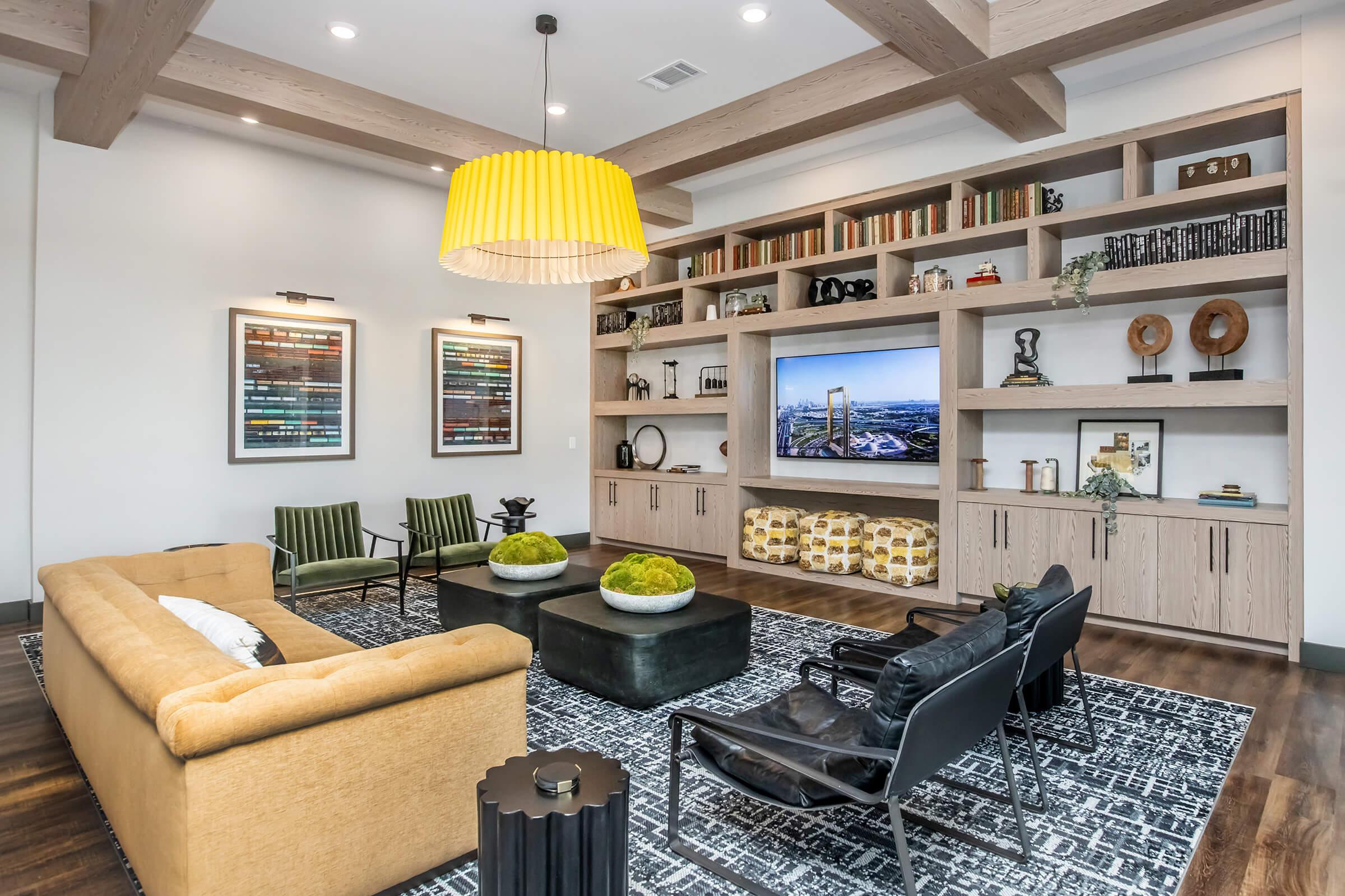 A modern living room featuring a yellow pendant lamp, a cozy sofa in a light tan color, and a dark coffee table. Black and green accent chairs are arranged near a stylish bookcase filled with decorative items and books. A large TV is mounted on the wall, enhancing the contemporary decor.