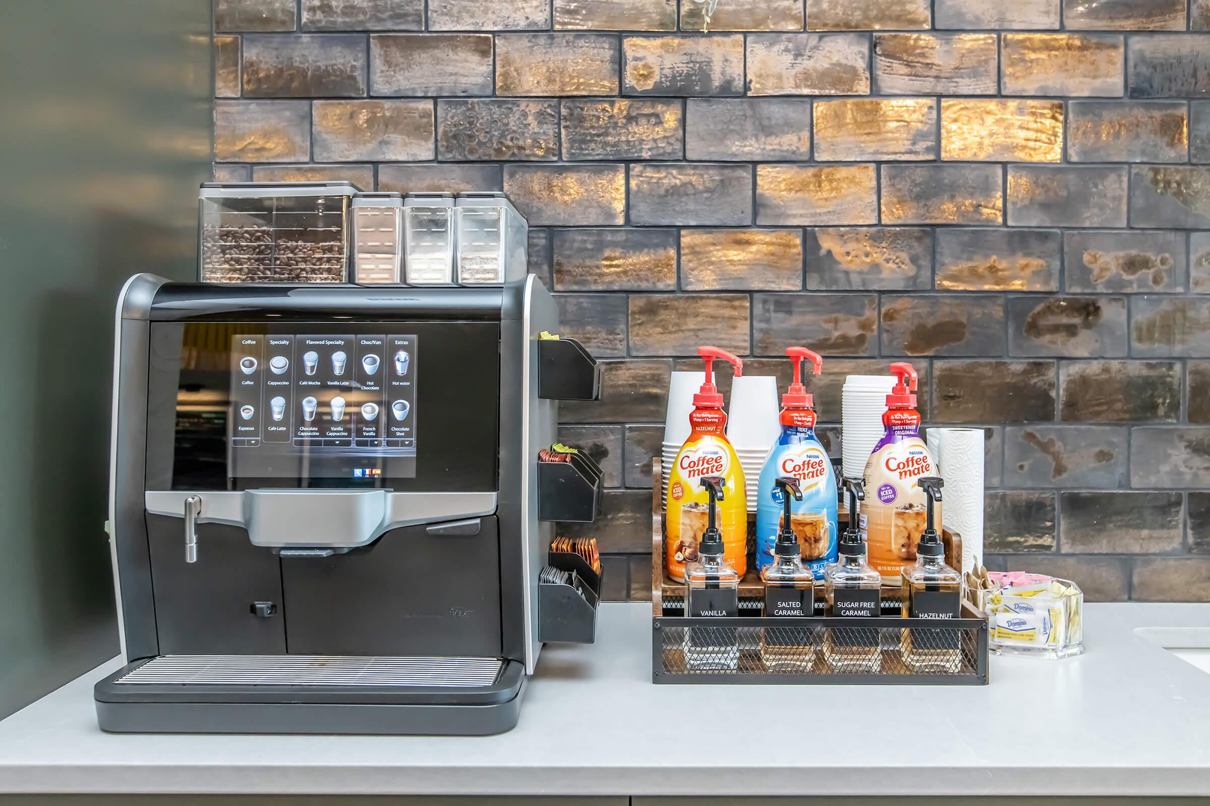 A modern coffee machine with a touchscreen interface sits on a countertop next to a variety of flavored coffee syrups in dispensers. Behind the machine, there is a textured wall of dark tiles. The setup suggests a stylish coffee station suitable for preparing beverages.