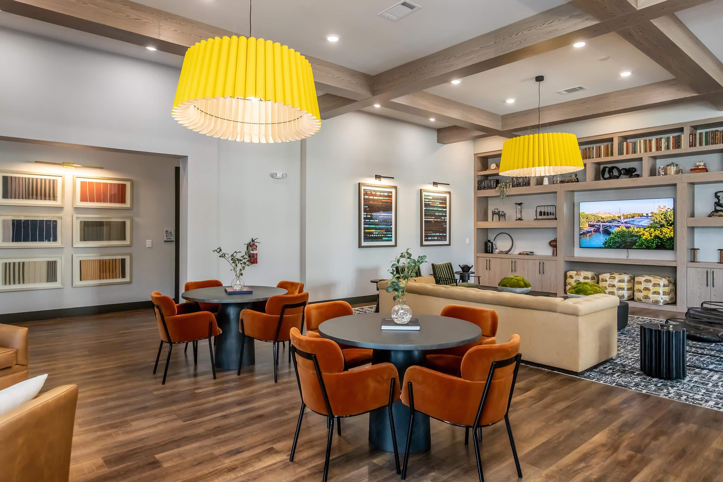 Modern living space featuring a cozy seating area with a beige sofa, two round tables surrounded by orange chairs, and a large entertainment unit with a TV. Bright yellow pendant lights hang from the ceiling, and there are artworks on the wall along with decorative items on shelves. Natural light enhances the inviting atmosphere.