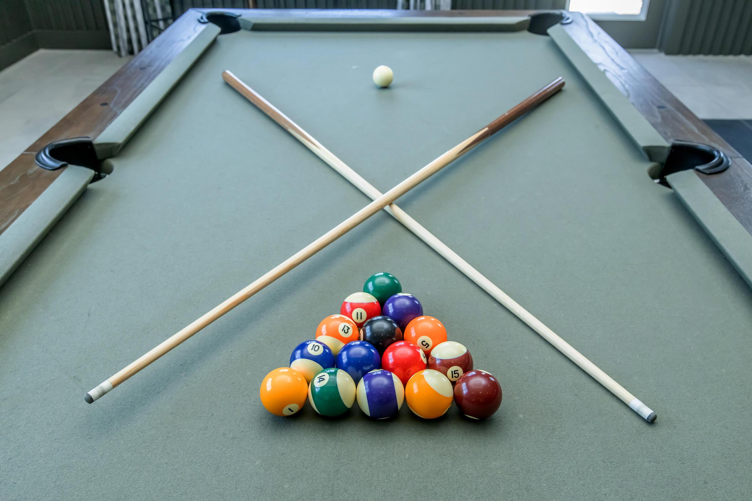 A billiards table with a triangle of colored pool balls arranged at the base, featuring a cue ball on the table. Two pool cues are crossed above the triangle. The table has a green felt surface and is set in a well-lit room, with a window visible in the background.
