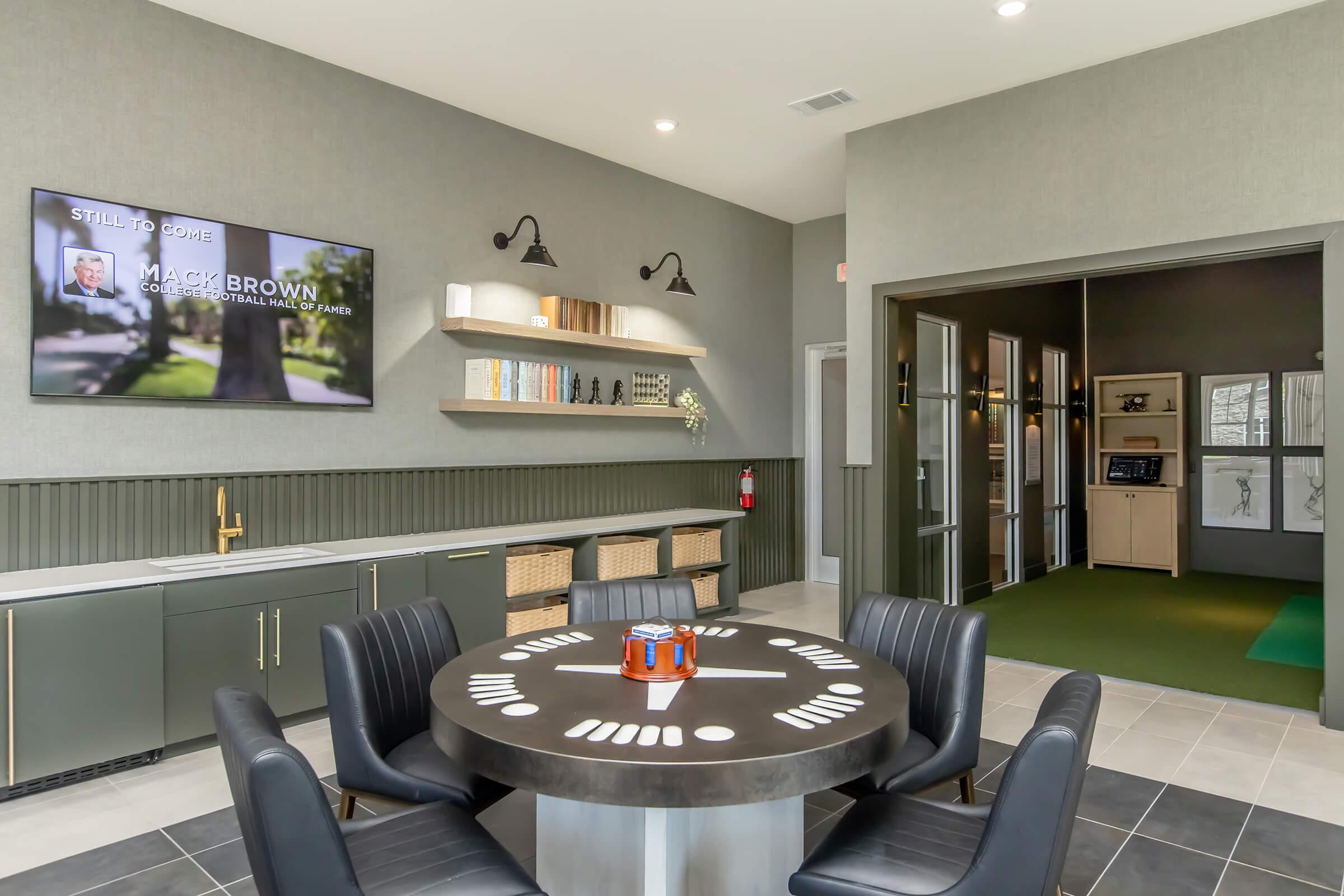 Modern interior of a shared lounge area featuring a round table with seating, a wall-mounted TV displaying a name, and stylish shelves with decorative items. To the right, there’s an entrance leading to a green space, possibly for activities or golf. The color scheme is a mix of gray and green tones.