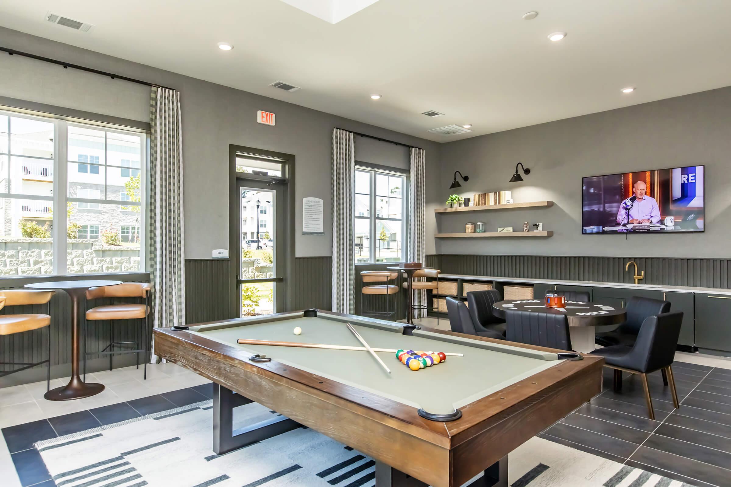A modern lounge area featuring a pool table in the center, surrounded by stylish seating arrangements. Large windows provide natural light, and a wall-mounted TV displays a show. The decor includes black and gray tones with accents of wood, creating a contemporary and inviting atmosphere.