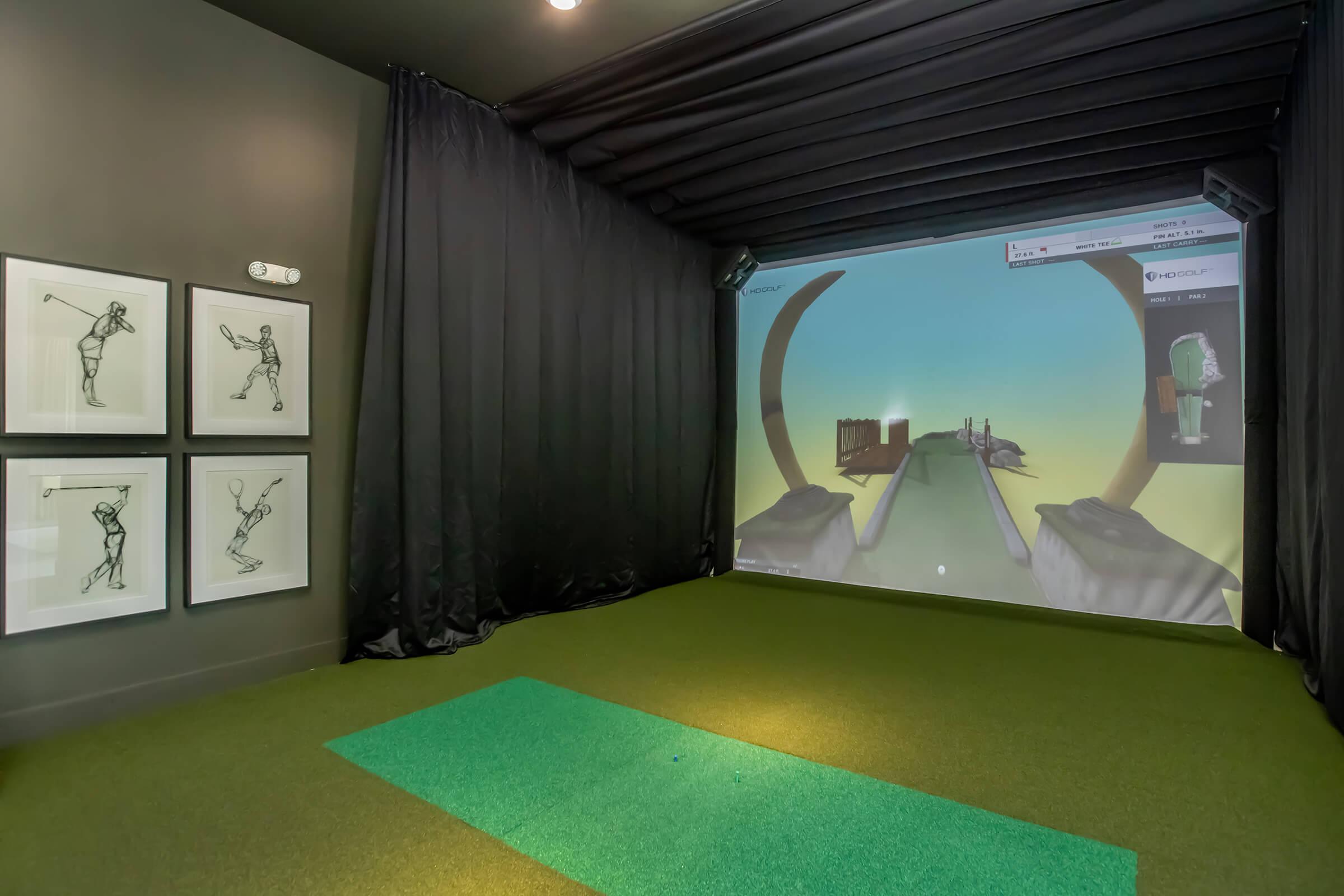 A virtual golf simulator setup in a dimly lit room. The image features a large projection screen displaying a golf course, with a green artificial turf mat in the foreground. Black curtains surround the area, and framed illustrations of golfers are mounted on the wall.