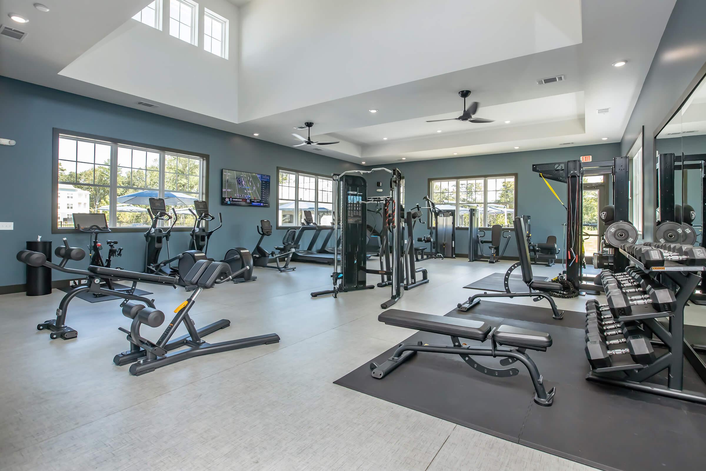 A modern gym interior featuring various fitness equipment, including weight benches, machines, and cardio machines. Large windows provide natural light, and the space has a sleek design with gray and blue tones. Two ceiling fans and televisions are visible, creating a welcoming workout environment.