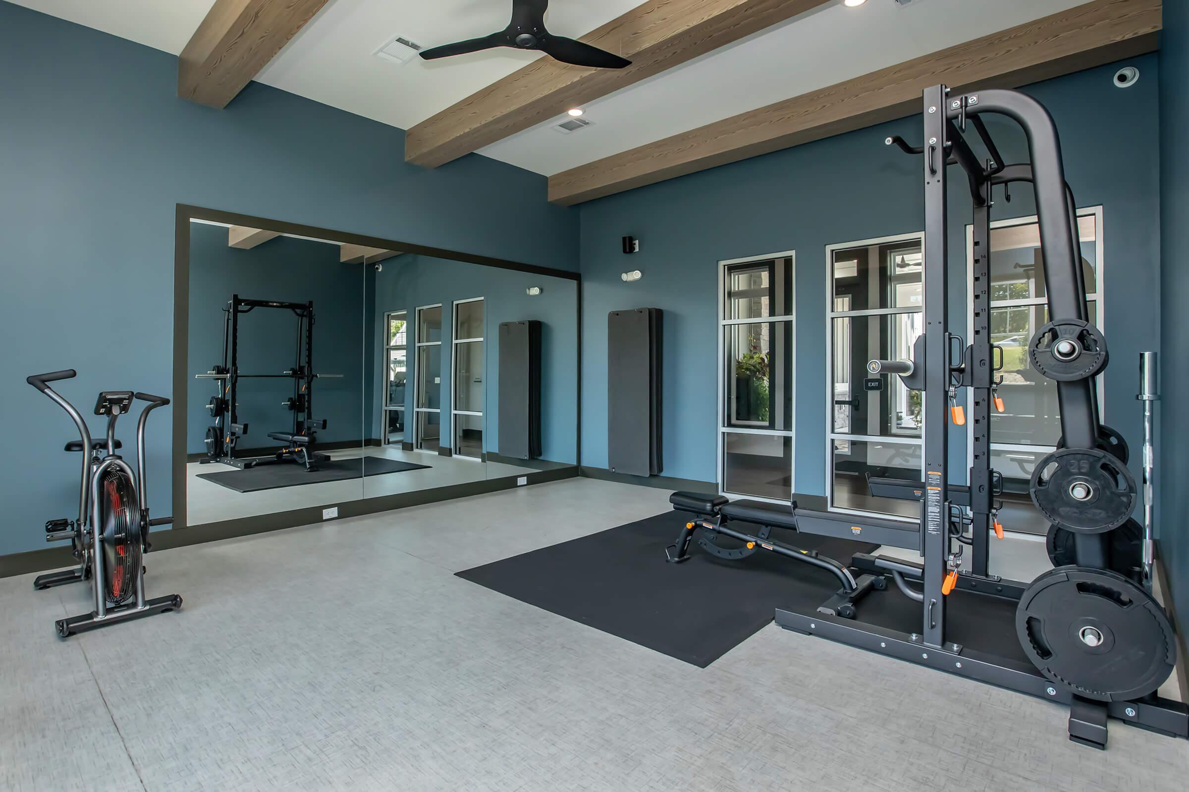 A modern home gym with a mirrored wall, featuring a multifunctional weight machine, an exercise bike, free weights, and a yoga mat on the floor. The room has neutral-colored walls with wooden beams and large windows for natural light.