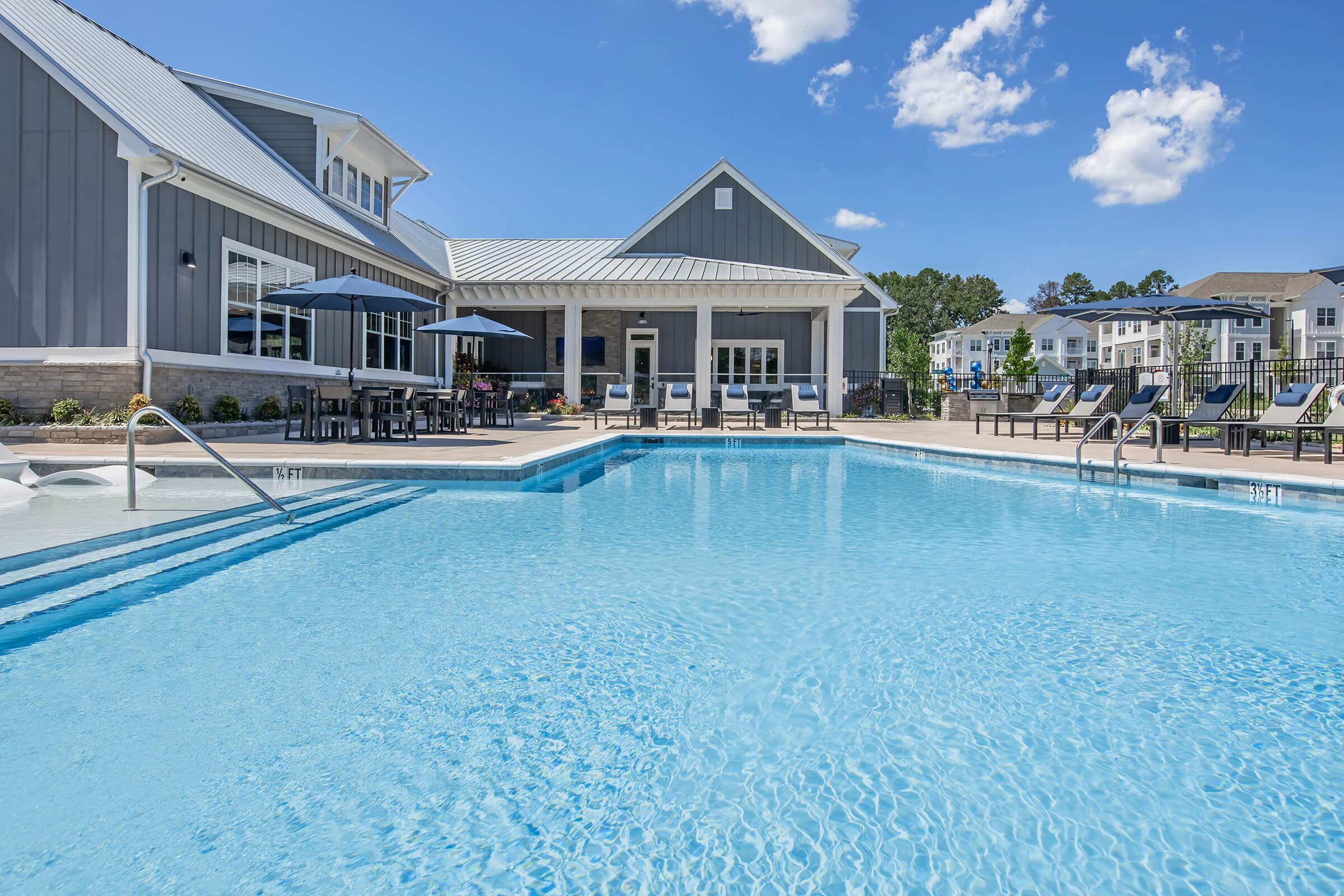 A bright and inviting outdoor swimming pool with clear blue water, surrounded by lounge chairs and umbrellas. In the background, there's a contemporary clubhouse with a covered patio, and well-maintained landscaping under a clear blue sky dotted with a few fluffy clouds.