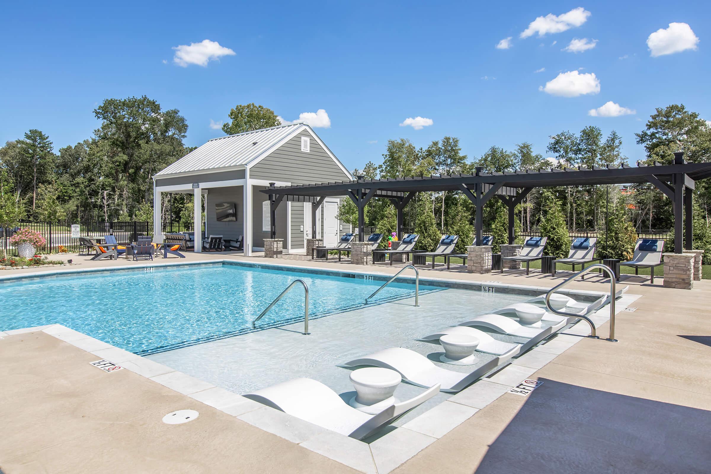 A sunny outdoor pool area featuring a clear blue pool with lounge chairs around it. There is a shaded patio structure and a small building in the background. Lush greenery and trees are visible, creating a relaxing atmosphere.
