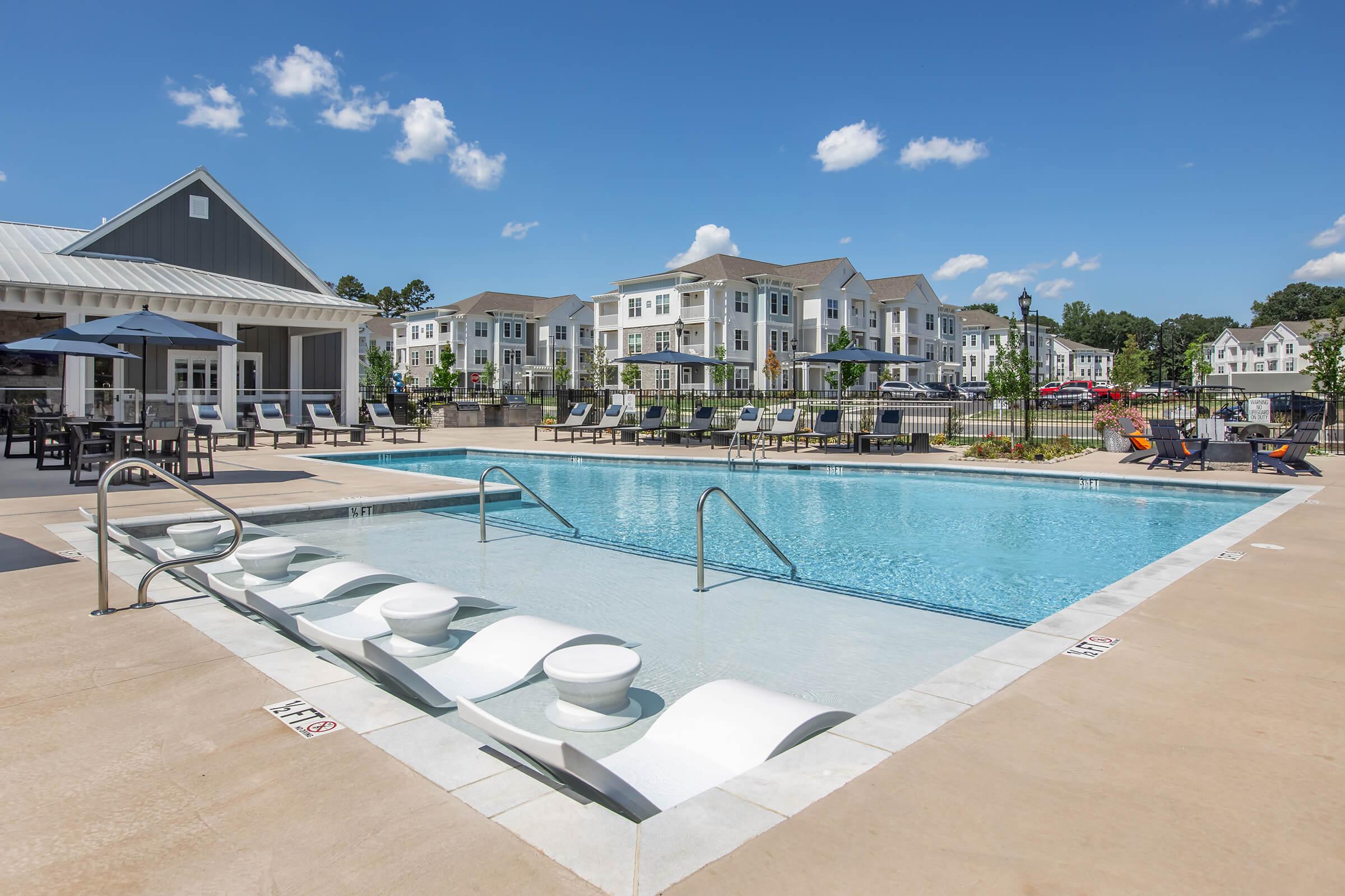 A spacious outdoor swimming pool featuring a shallow entry area, surrounded by lounge chairs and umbrellas. In the background, modern apartment buildings with a clear blue sky and fluffy white clouds. The scene conveys a relaxing, resort-style atmosphere.