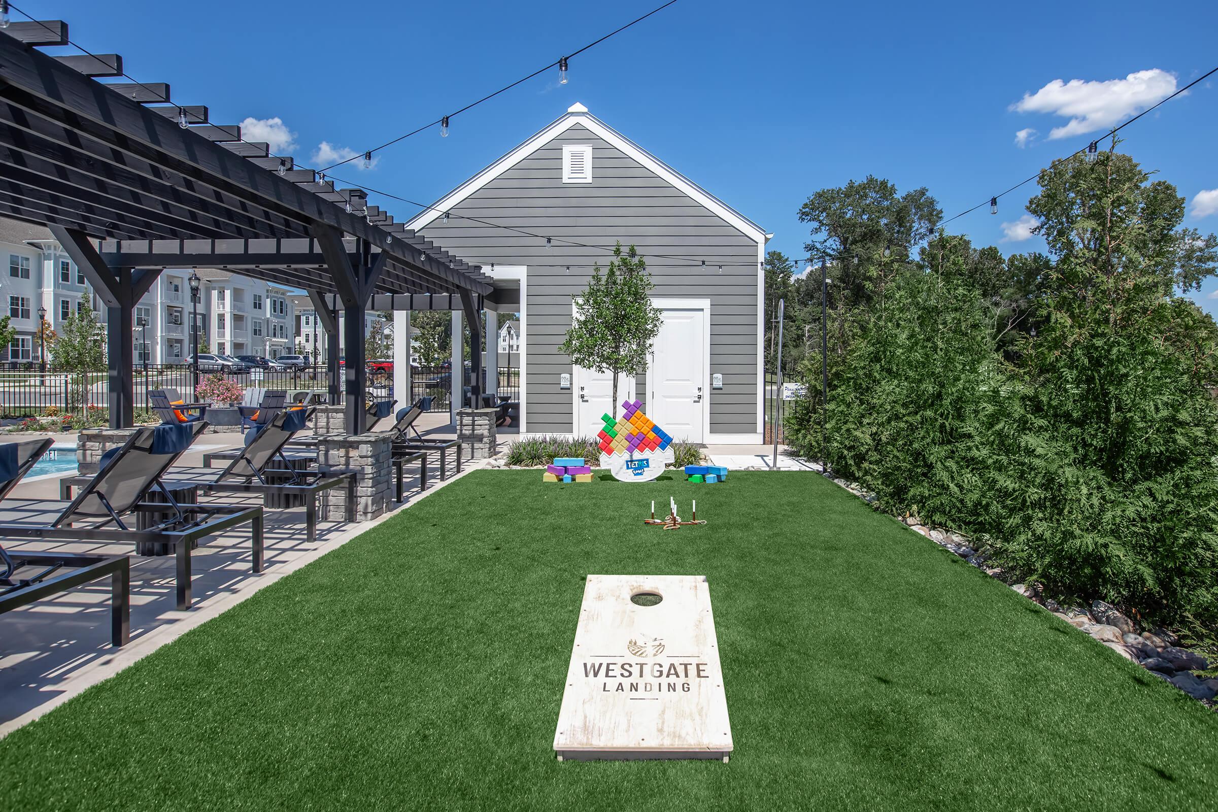 A vibrant outdoor area featuring a well-maintained lawn, a cornhole game with a branded board, and colorful yard games. There are lounge chairs and a pergola providing shade, with a modern gray building in the background. Strings of lights add a festive touch to the space, ideal for gatherings and relaxation.