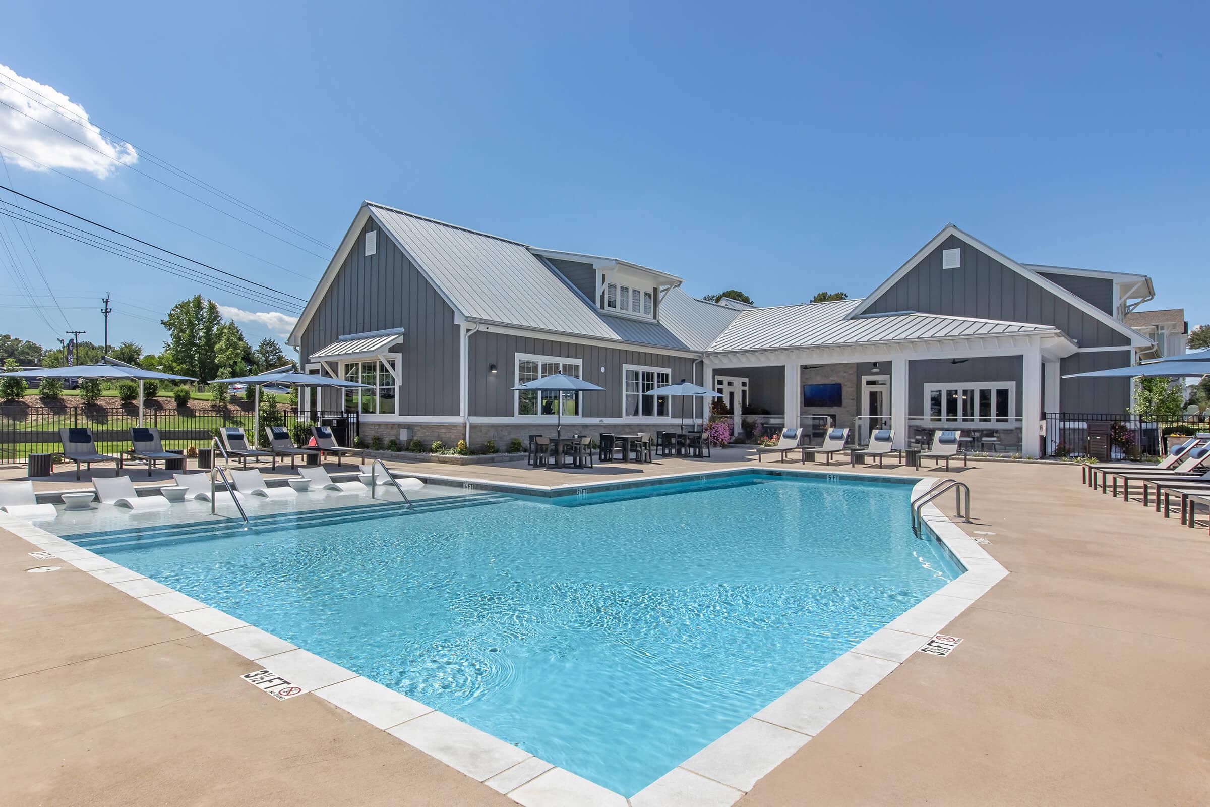 A modern outdoor swimming pool with lounge chairs and umbrellas, set against a clear blue sky. In the background, there is a stylish building featuring large windows and a gray exterior. Green trees line the horizon, enhancing the tranquil atmosphere of the residential area.