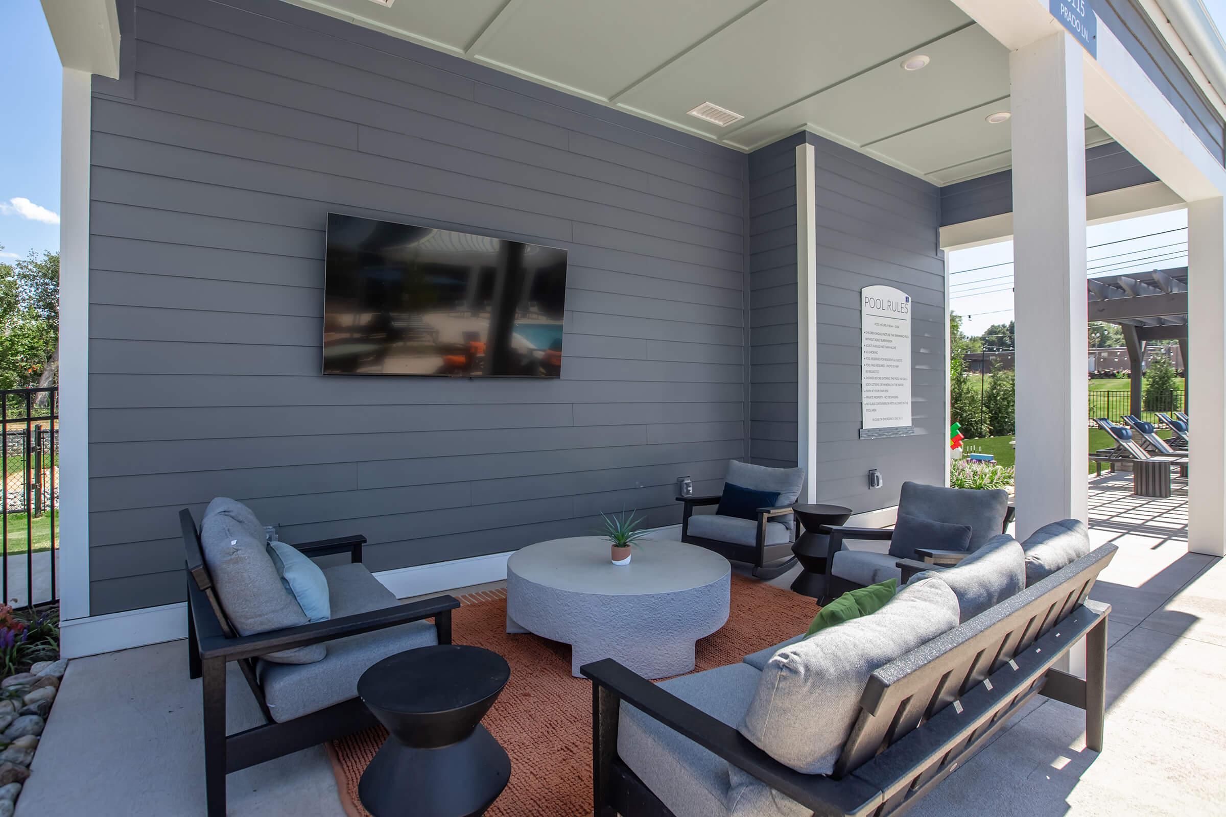 A modern outdoor lounge area featuring comfortable seating on a patio, a circular coffee table, a wall-mounted TV, decorative plants, and a bright blue sky in the background. Ideal for relaxation and social gatherings.