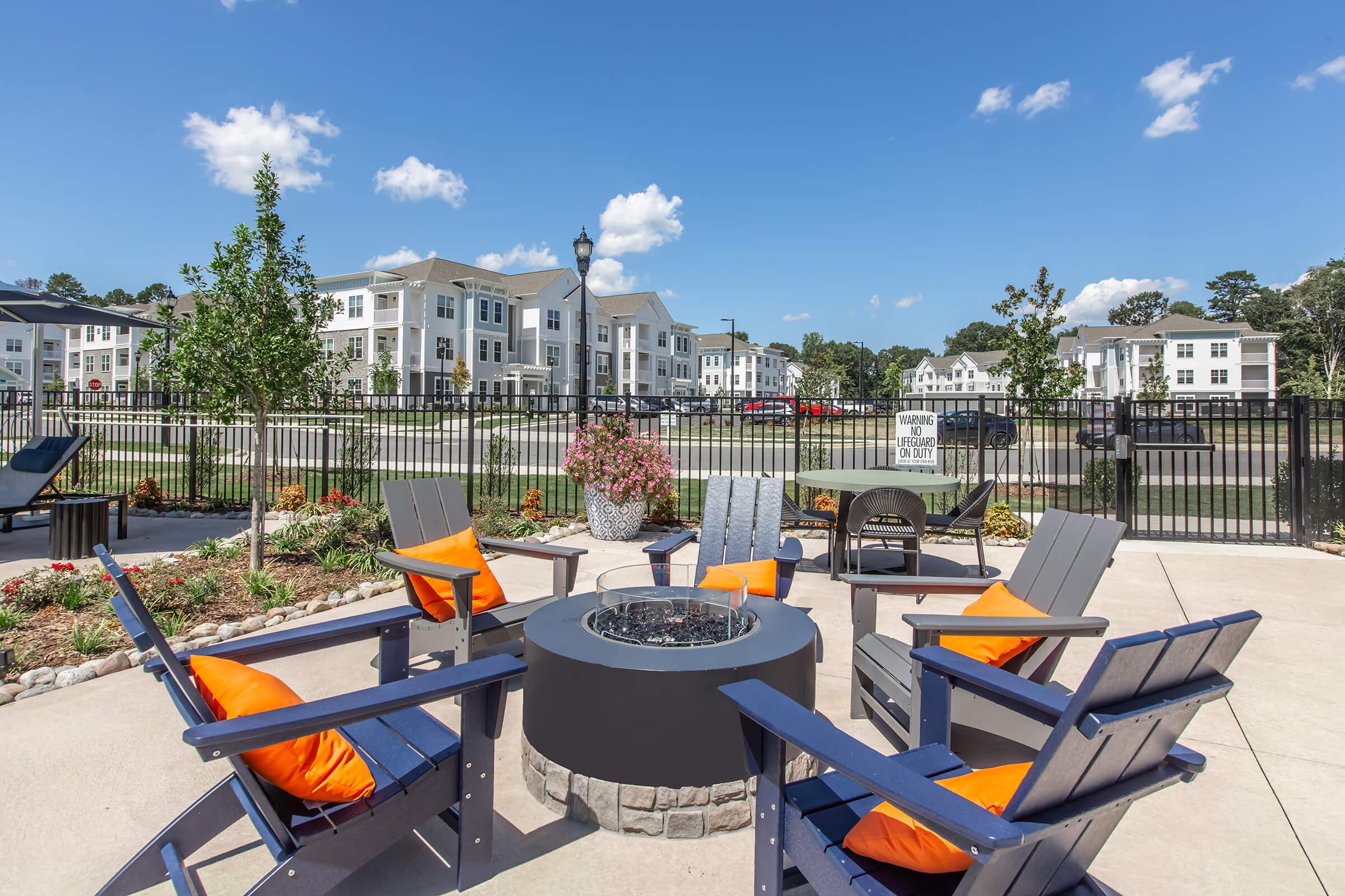 Outdoor seating area with blue adirondack chairs and orange cushions surrounding a fire pit, landscaped with flowers and trees. In the background, modern apartment buildings are visible under a clear blue sky with a few clouds.
